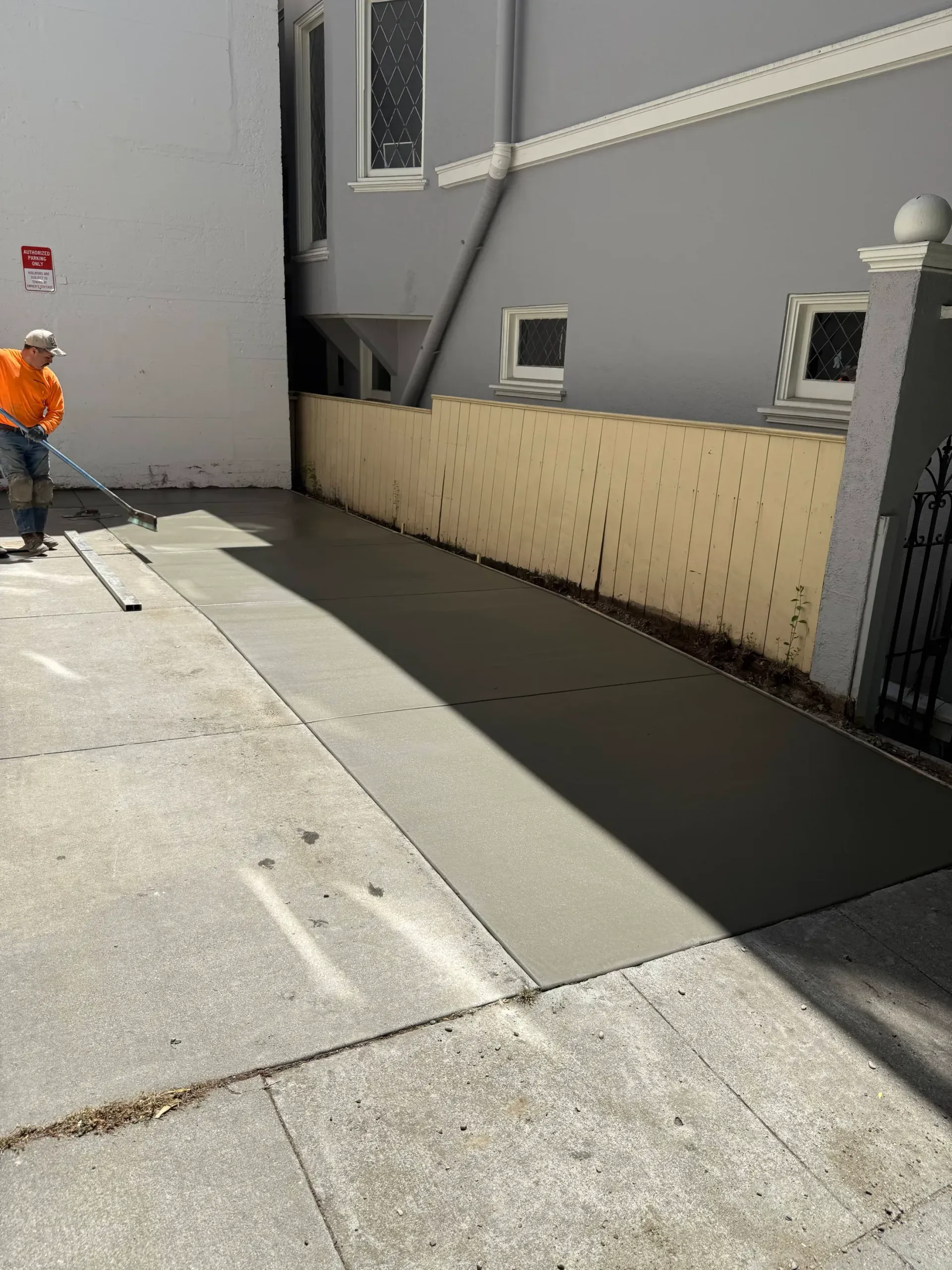 Man smoothing wet concrete in alleyway. Gray concrete surface, light beige wall, building on either side.