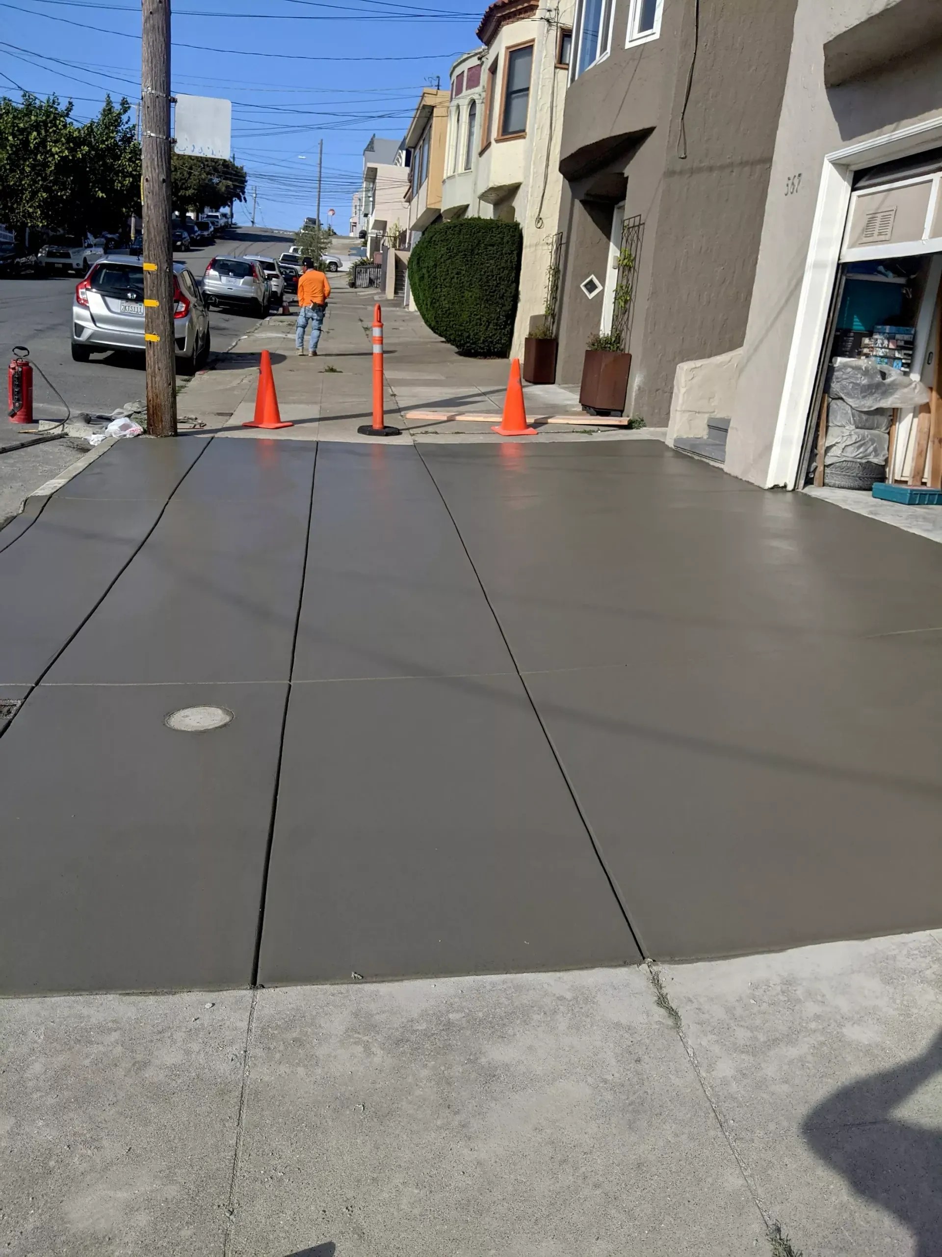 Freshly poured concrete sidewalk with orange traffic cones and a utility worker in the background on a city street.