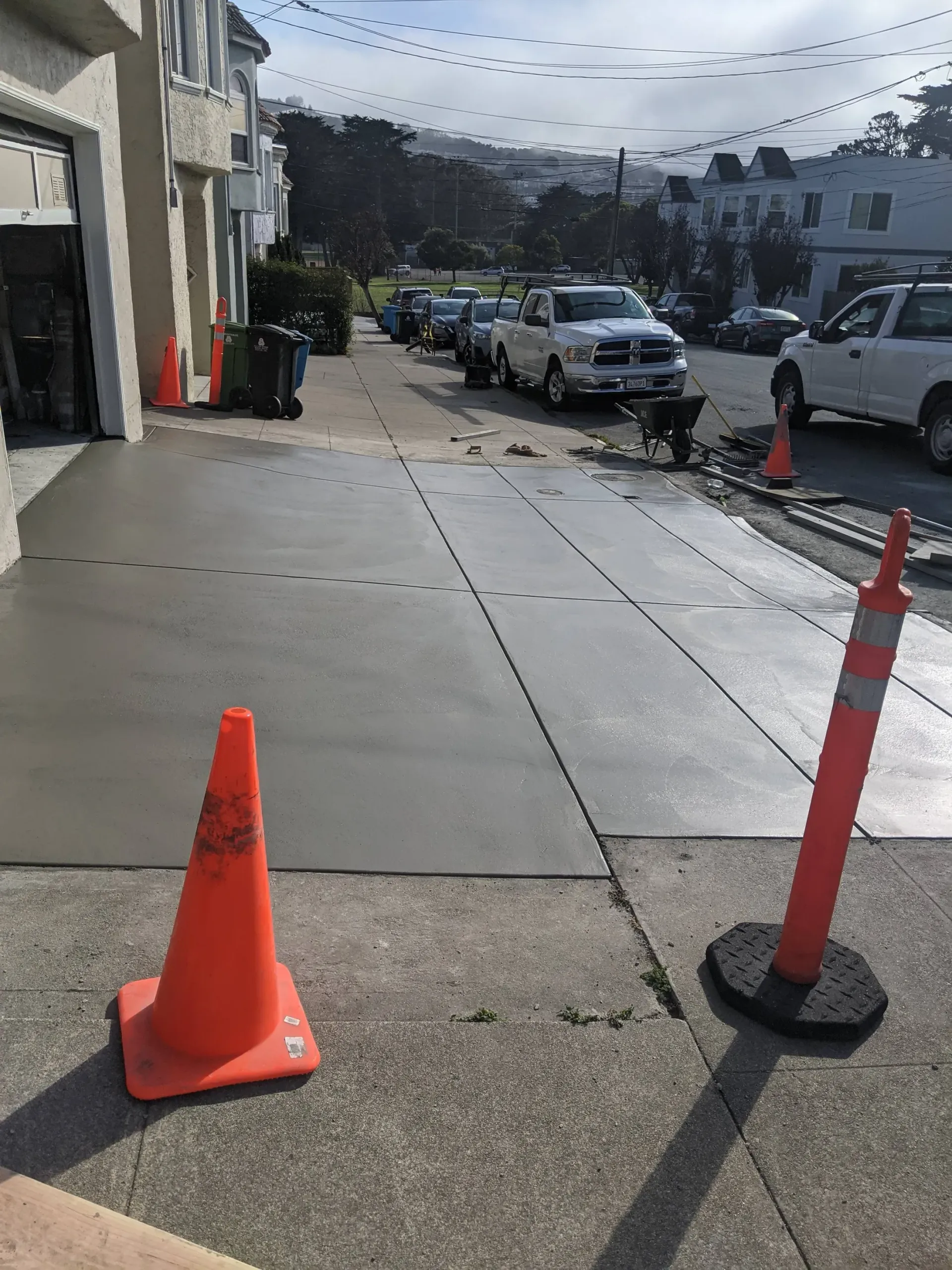 Freshly poured concrete driveway with orange safety cones; several parked vehicles in the background.