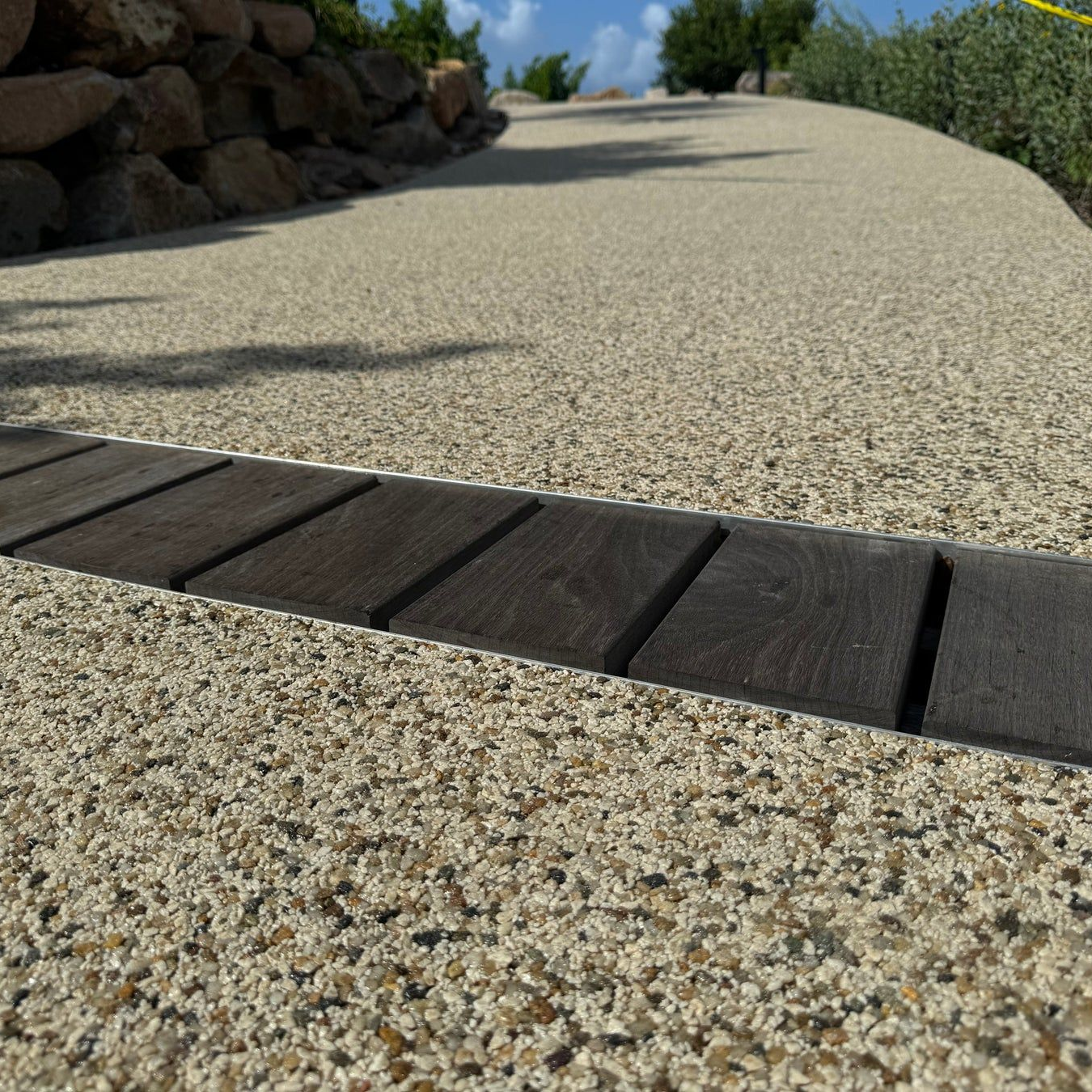Beige speckled pathway with dark wood slats, leading uphill towards green vegetation and blue sky.