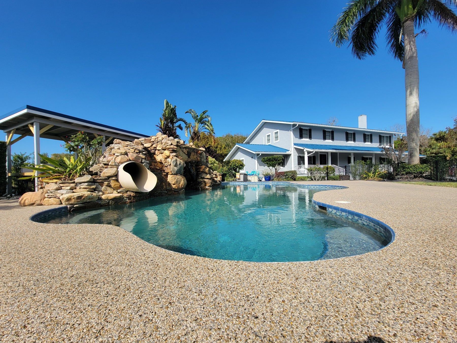 Pool with a waterfall slide and a light blue house under a clear, blue sky.