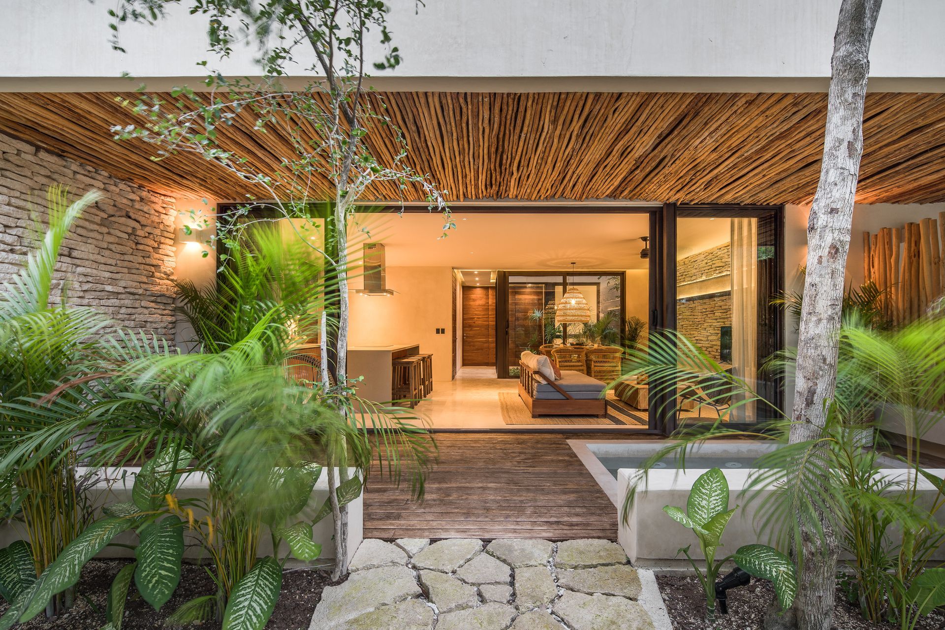 A house with a wooden ceiling and a sliding glass door is surrounded by trees and plants.