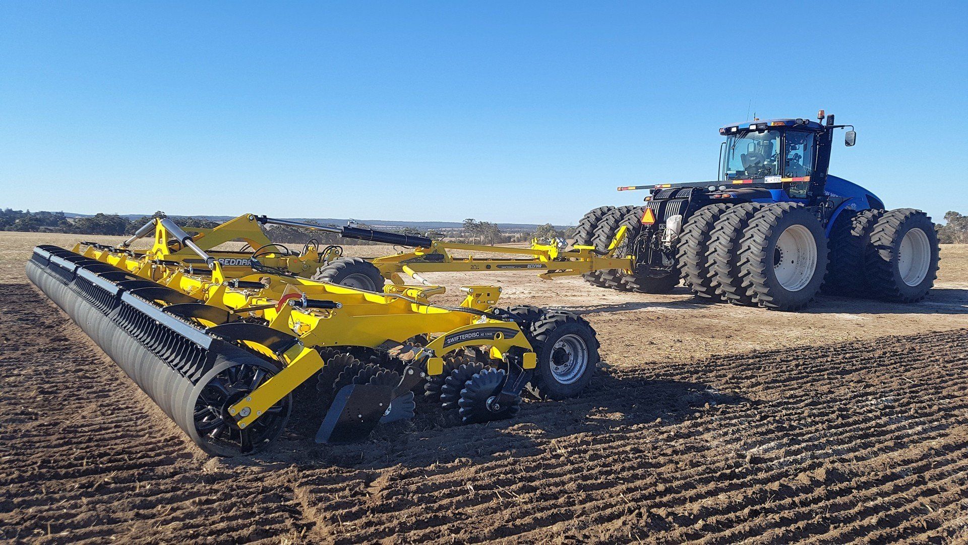 A large yellow roller towed by a blue tractor on a field, preparing the ground for planting under a clear sky.