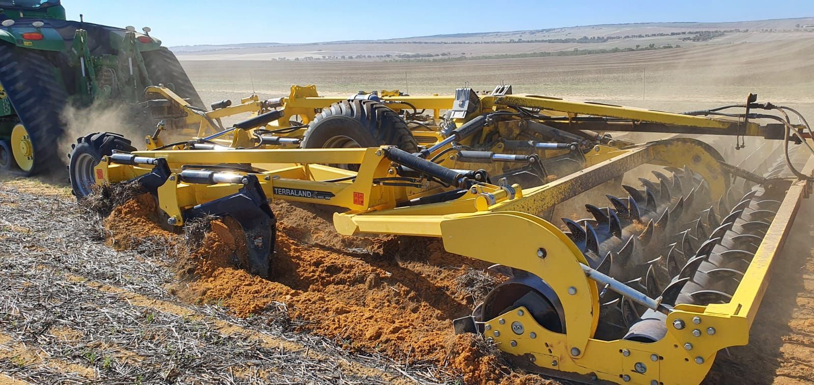 A large yellow agricultural roller being towed by a blue tractor on a dirt field under a clear blue sky.