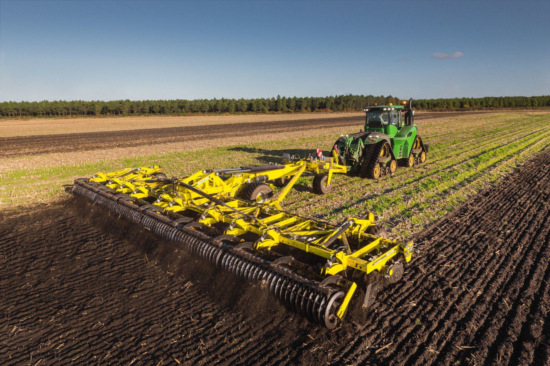 A green John Deere tractor pulling a yellow tillage implement across a field, under a clear blue sky.