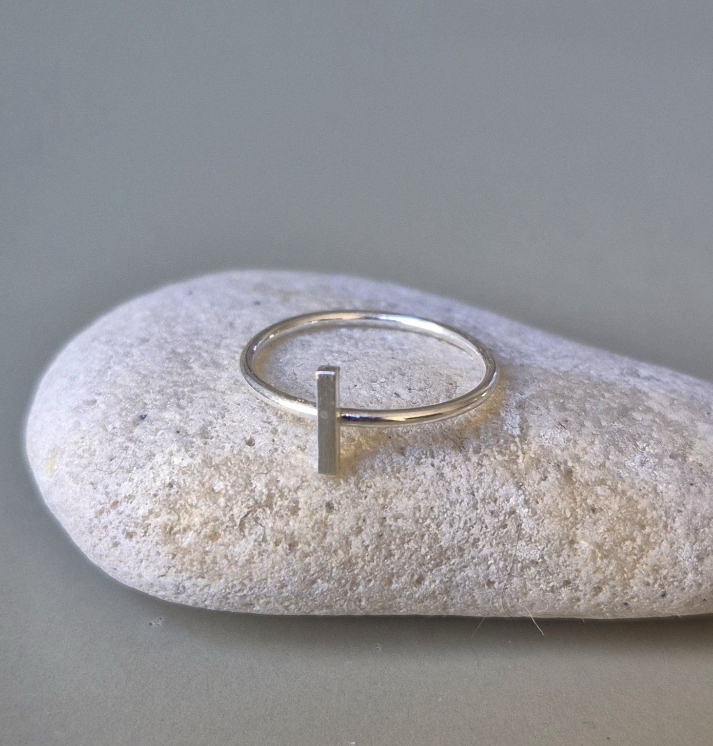 Anillo de plata con detalle de barra vertical sobre piedra lisa y gris.
