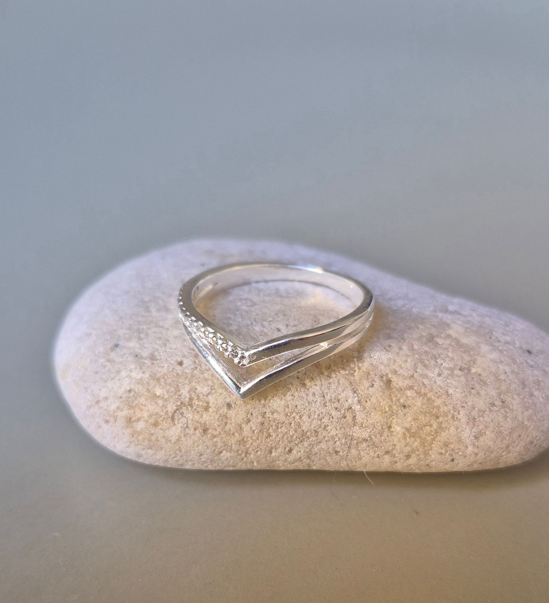 Anillo de plata en forma de chevron con pequeñas piedras, apoyado sobre una roca de color claro.