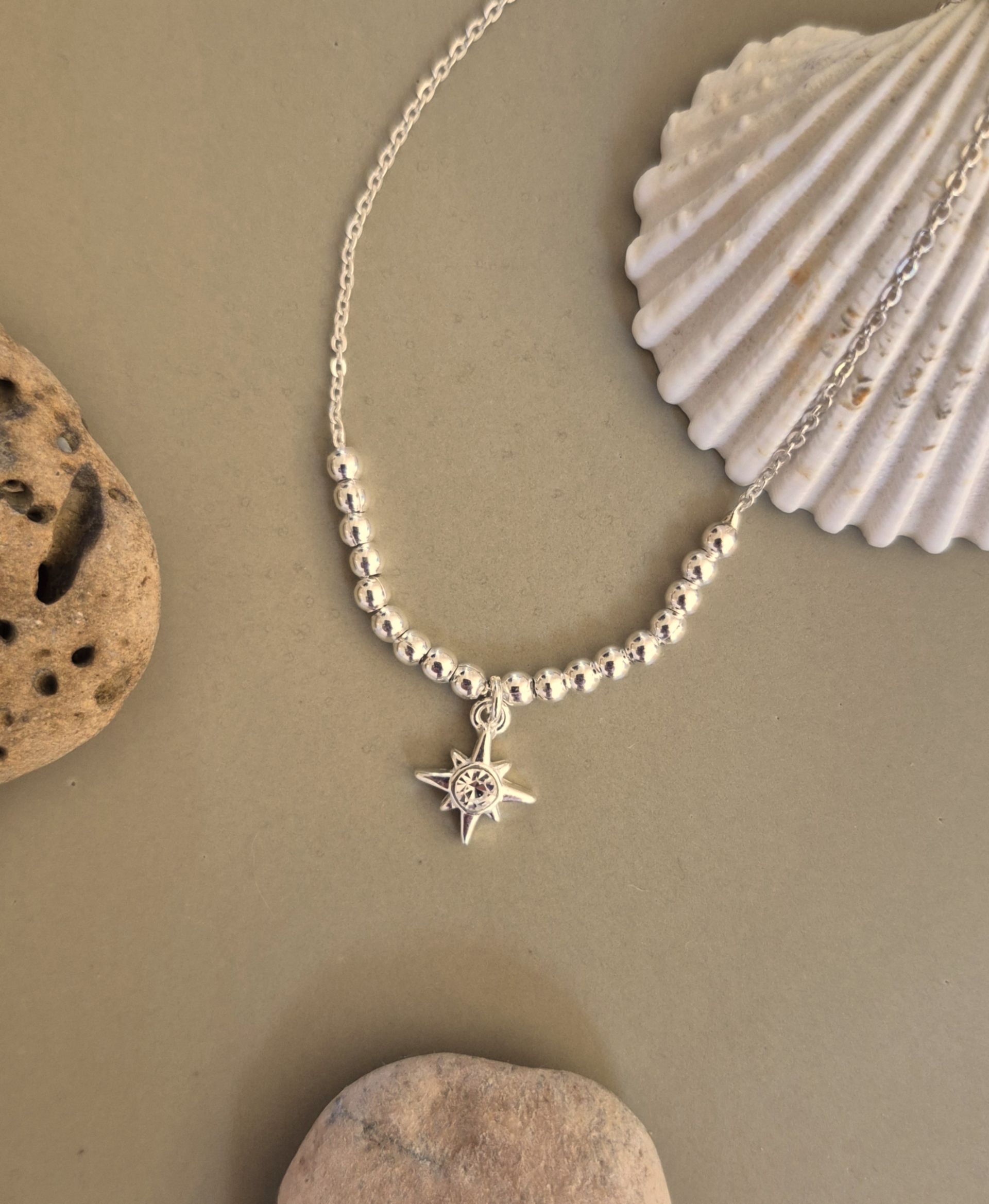 Collar de plata con colgante de estrella y cuentas. Superficie beige, con concha y piedras.