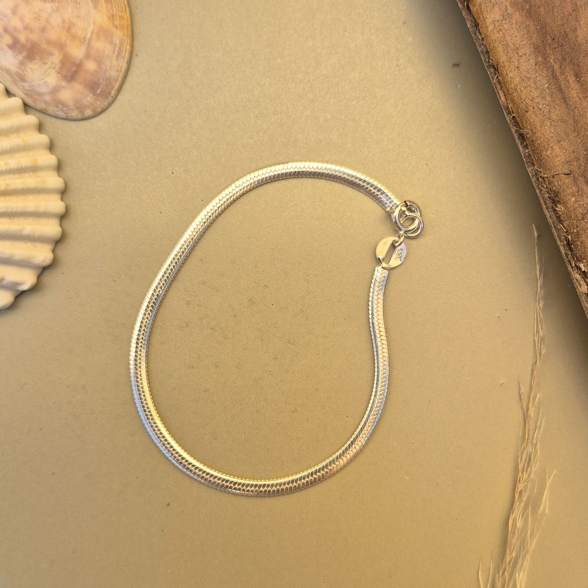 Pulsera de cadena de serpiente de plata sobre superficie beige, junto a una concha y madera.