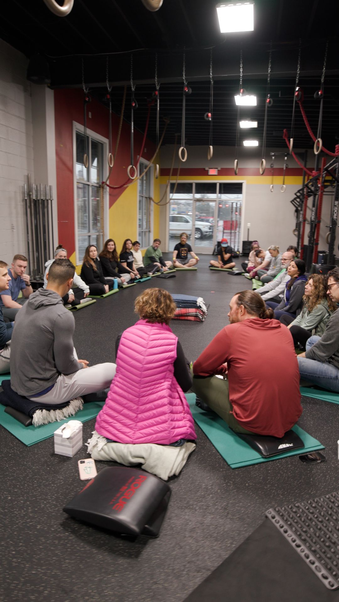 A group of people are sitting in a circle on yoga mats in a gym.