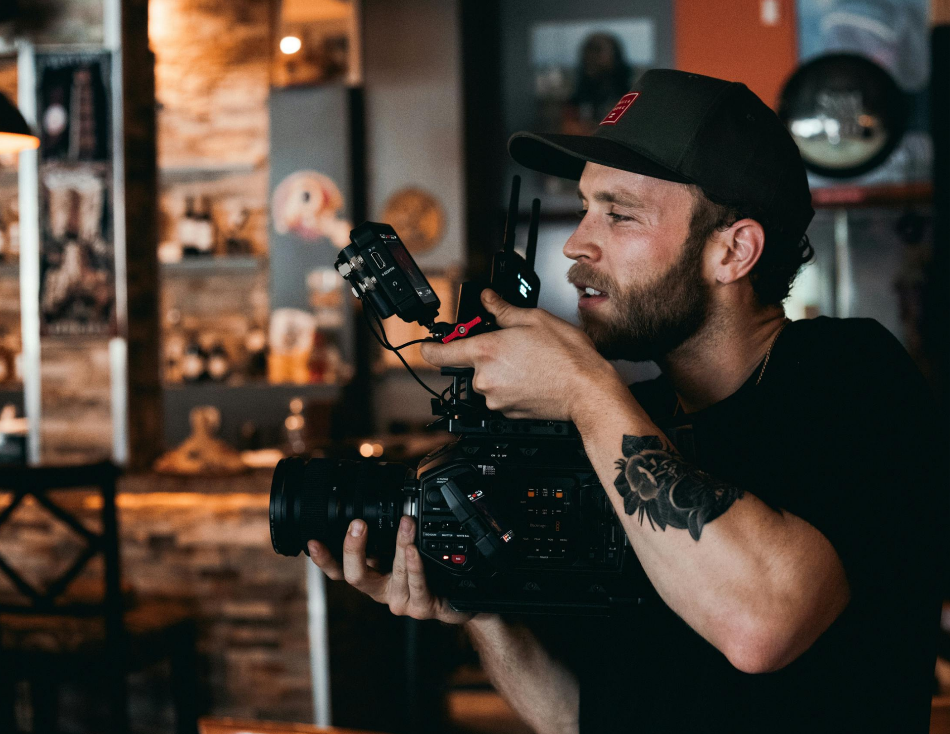 A bearded filmmaker wearing a dark baseball cap films with a professional cinema camera in a softly lit, rustic setting.