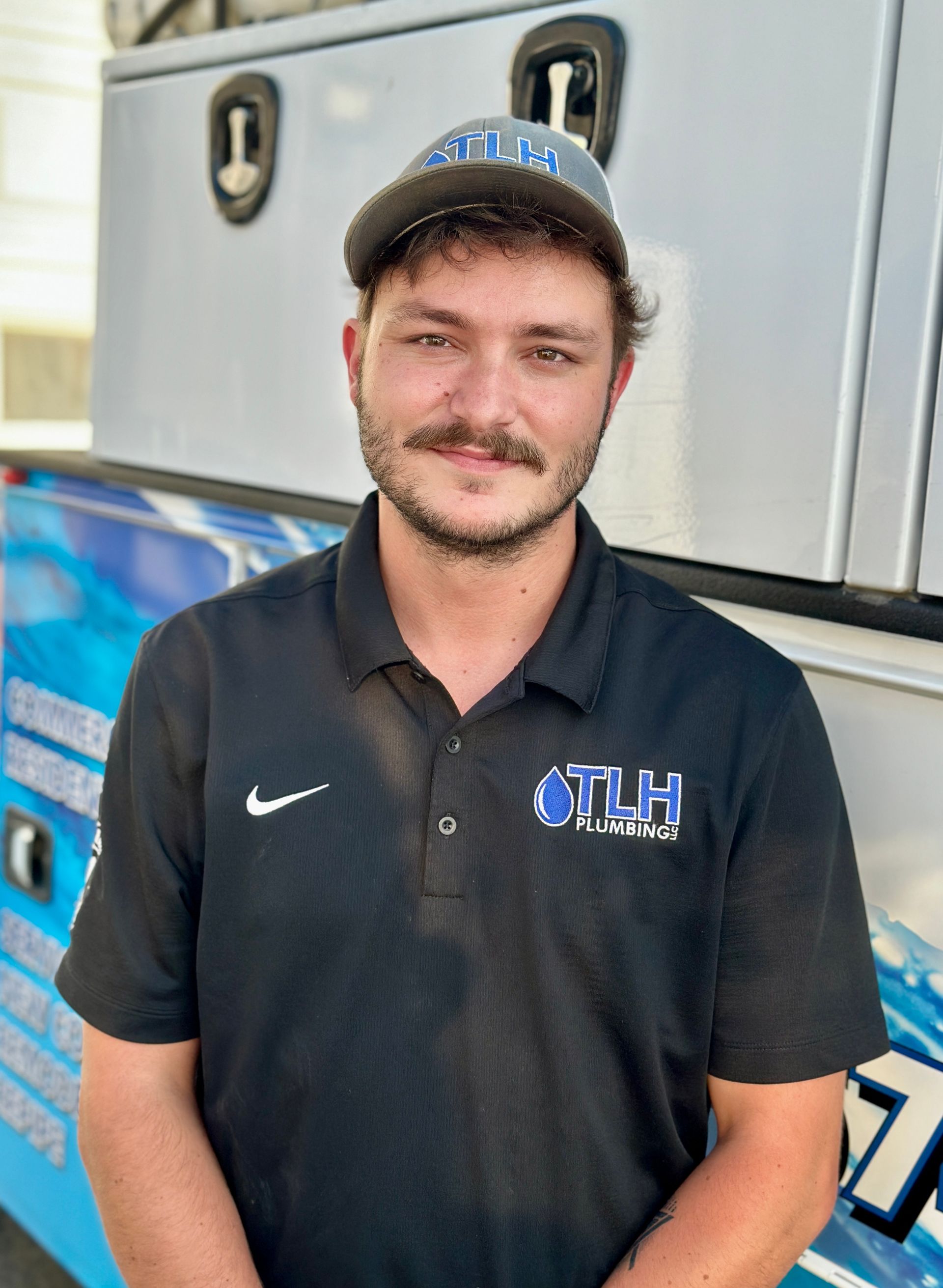 Man in black polo and cap stands near truck with logo.