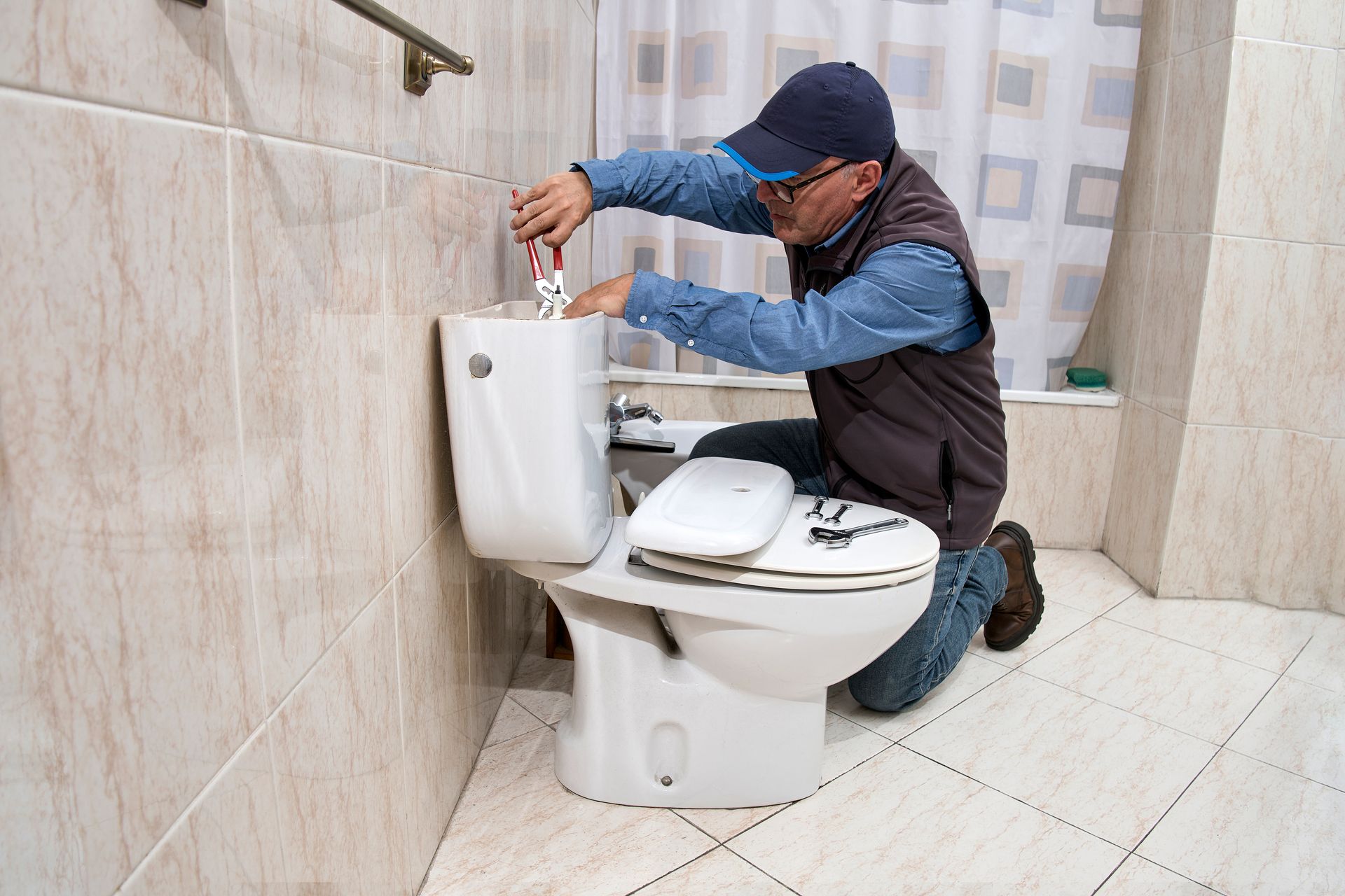Plumber repairing a toilet in a tiled bathroom, kneeling beside the tank and using a tool.