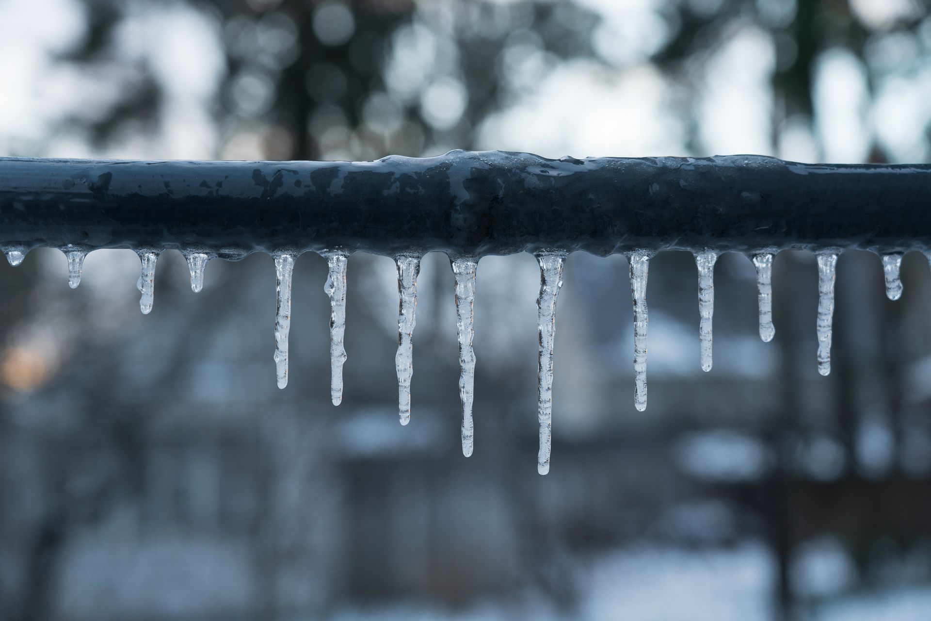 Icicles hang from a black pipe against a blurred, wintery background.