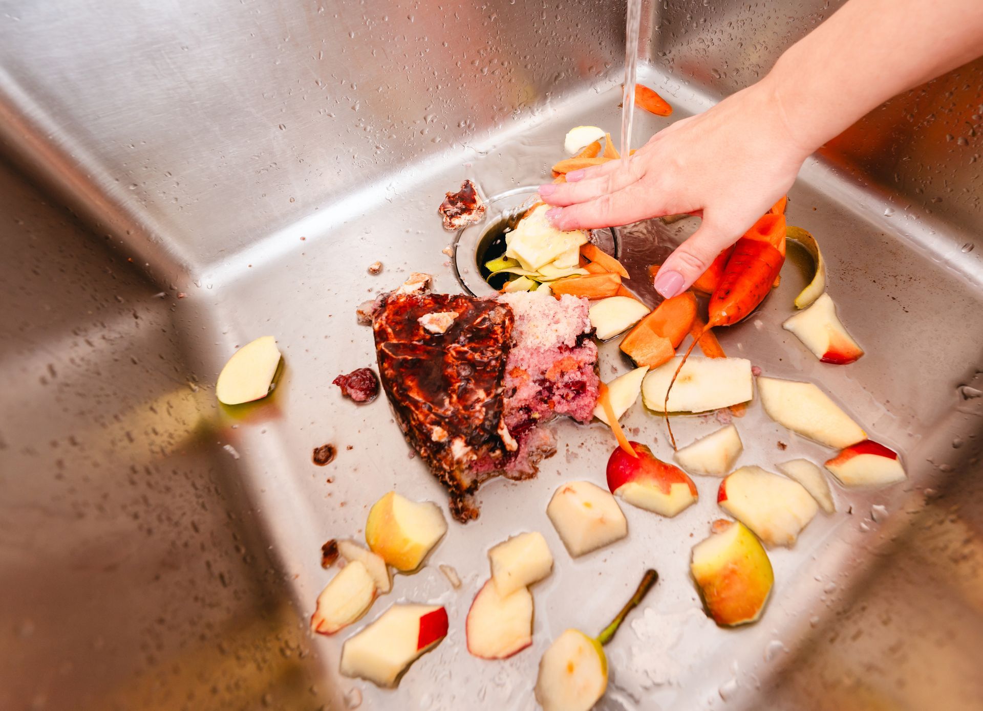 Hand rinsing food scraps, including fruit and meat, down a stainless steel kitchen sink drain.