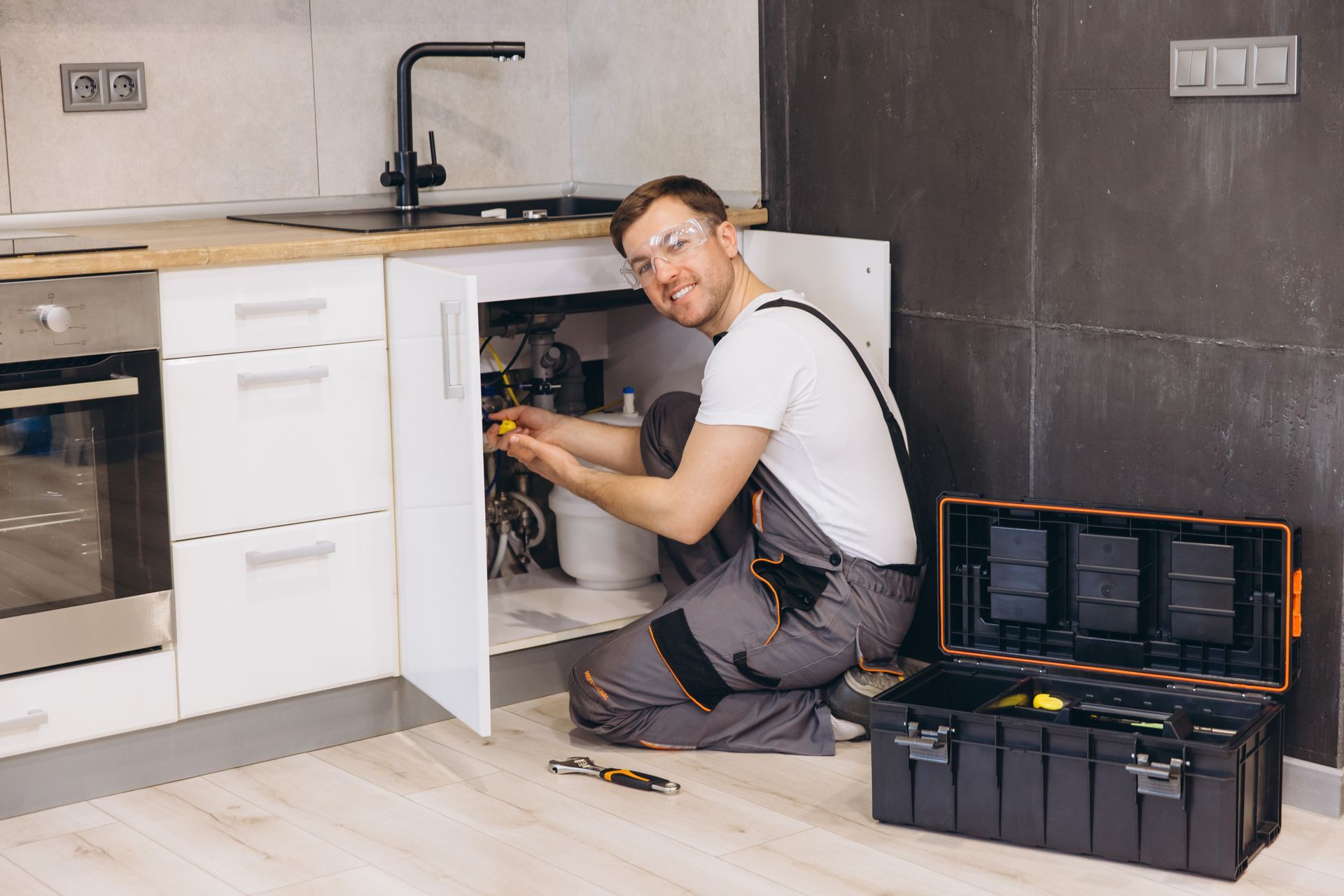 Plumber in overalls repairs kitchen sink with open toolbox, smiling, in front of white cabinets, dark gray walls.
