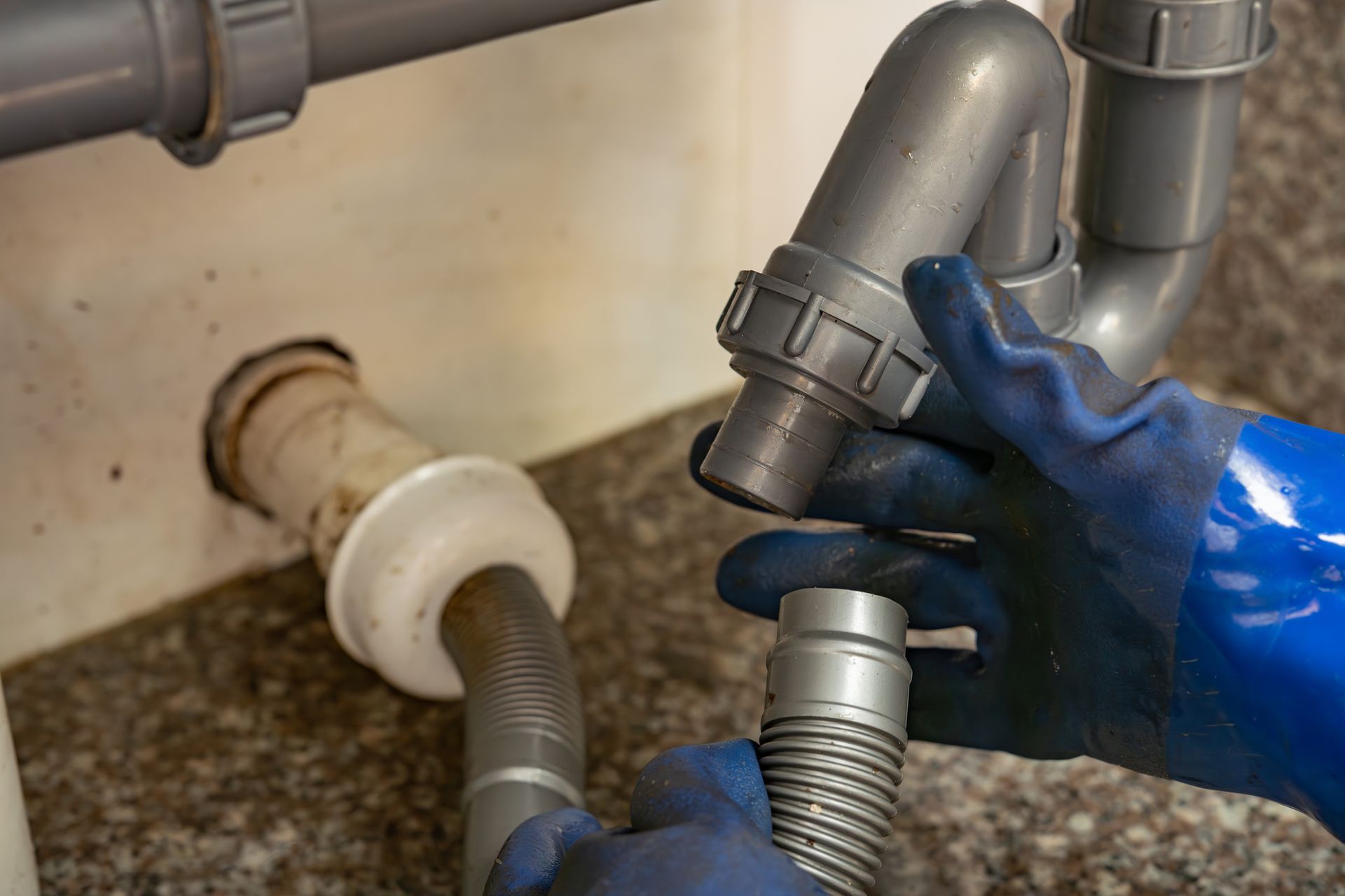 Hands in blue gloves connecting sink drain pipes under a sink.