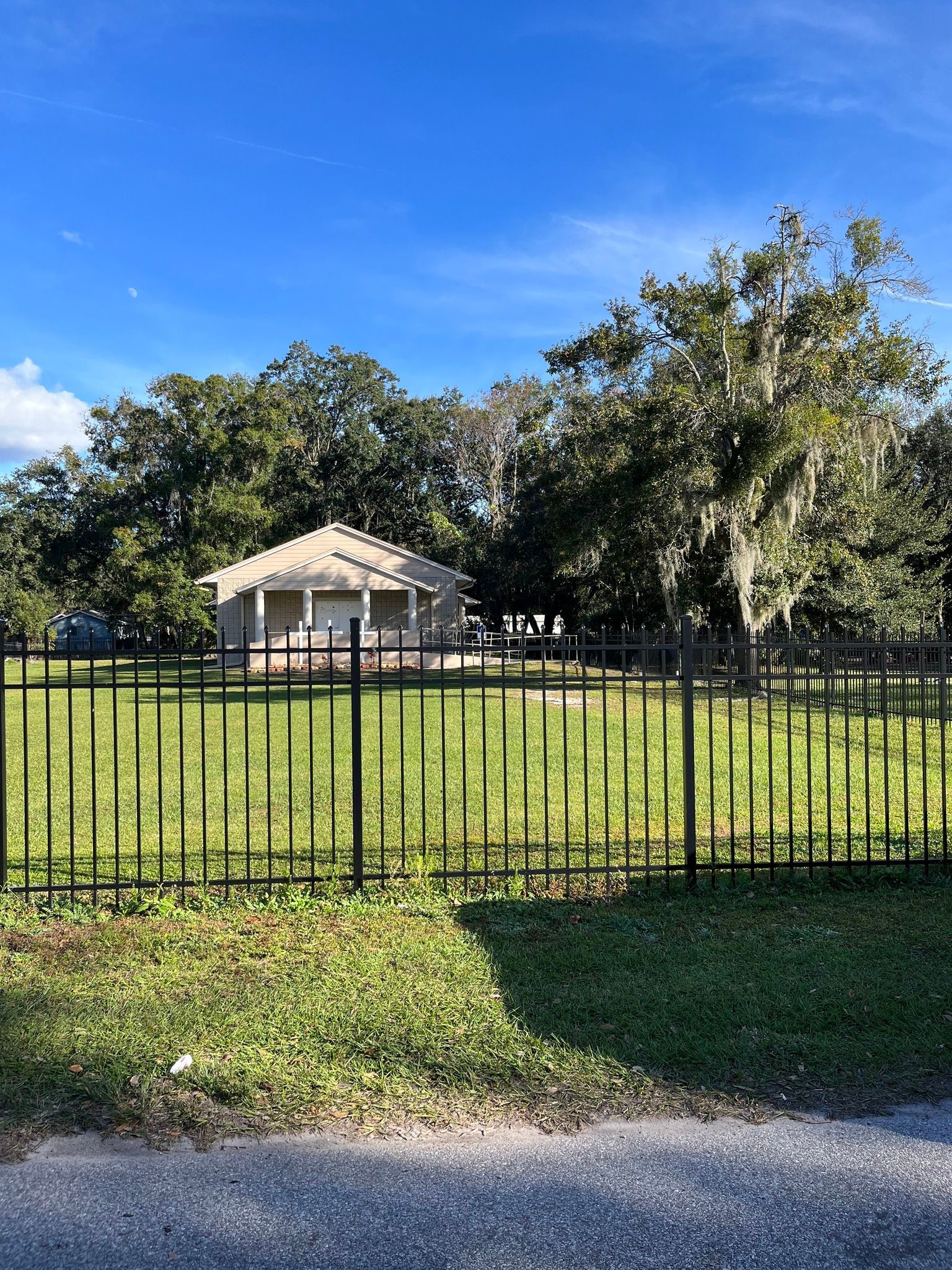 Small beige building behind a black metal fence, surrounded by green grass and trees, under a blue sky.