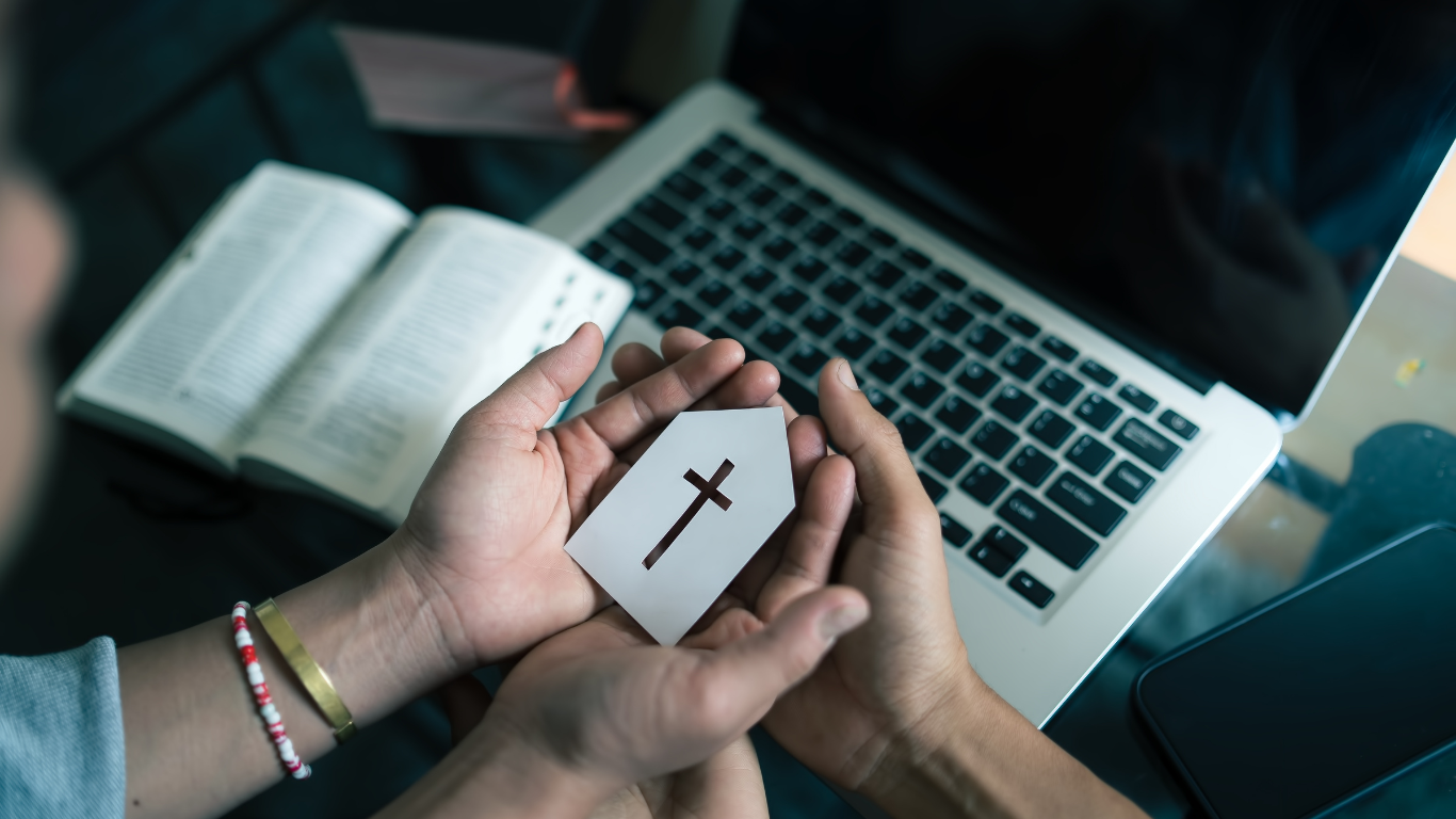 Hands holding a cross-shaped card over an open Bible and laptop.
