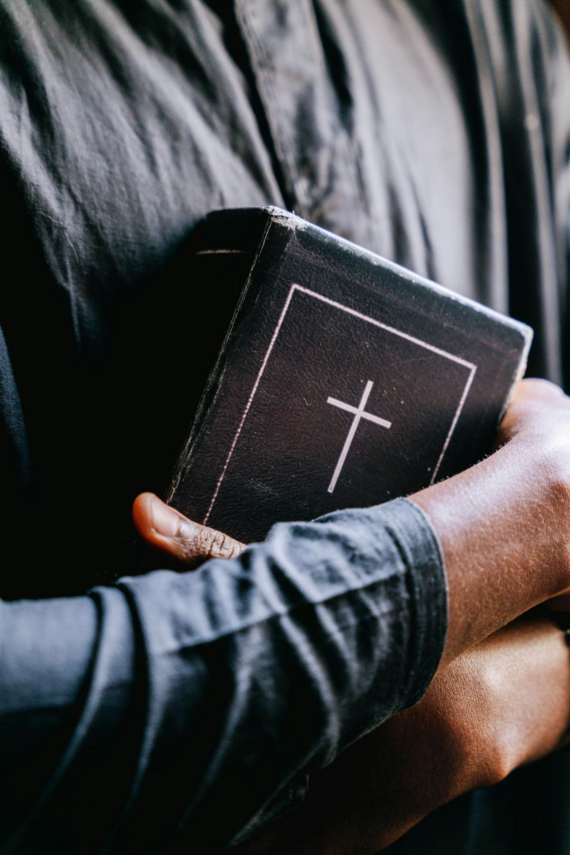 Person holding a Bible with a cross on the cover. Dark clothing.