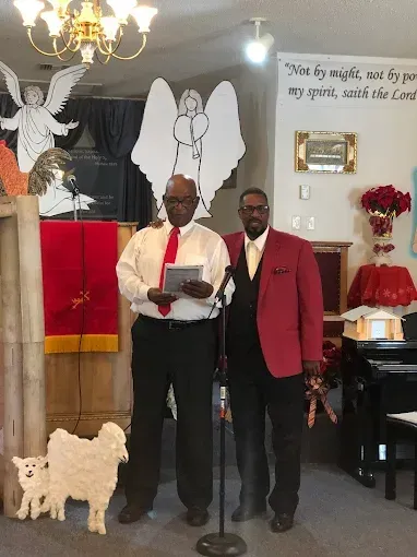 Two men standing near a microphone in a church; one holding a paper, red coat, white background.