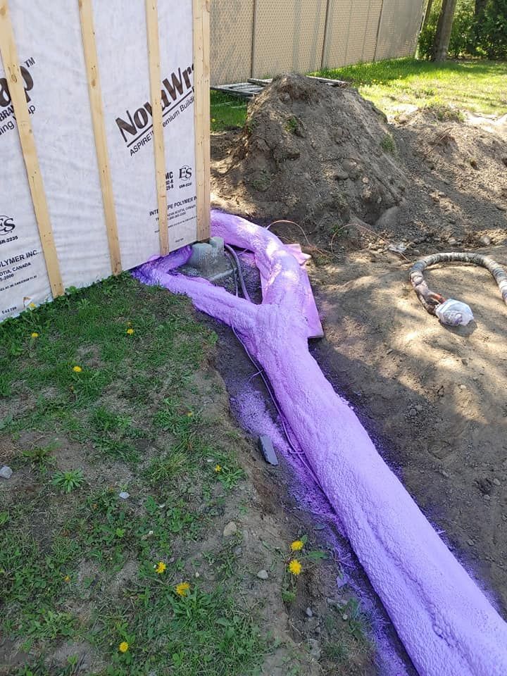 Isolation en mousse violette pulvérisée autour d'une fondation contre un mur à ossature de bois, sur de l'herbe et de la terre.