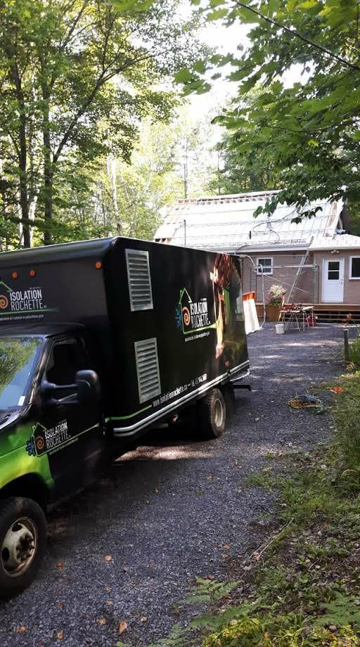 Camion stationné dans une allée de gravier devant une maison, avec une réparation de toit en cours, entouré d'arbres.