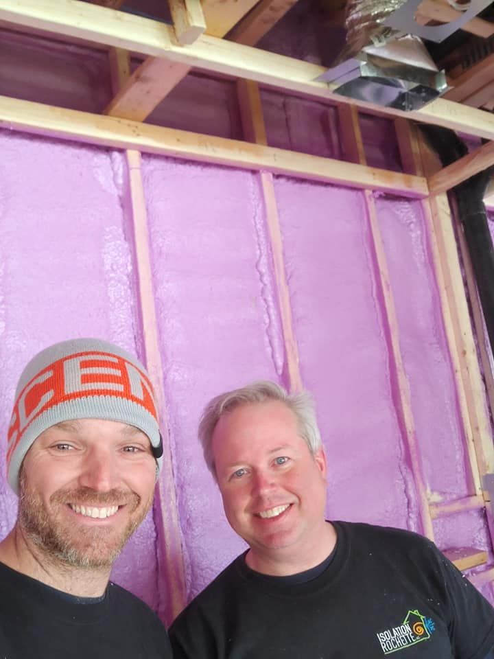Deux hommes souriant devant une isolation en mousse pulvérisée rose dans une pièce partiellement construite, avec des poutres en bois visibles.