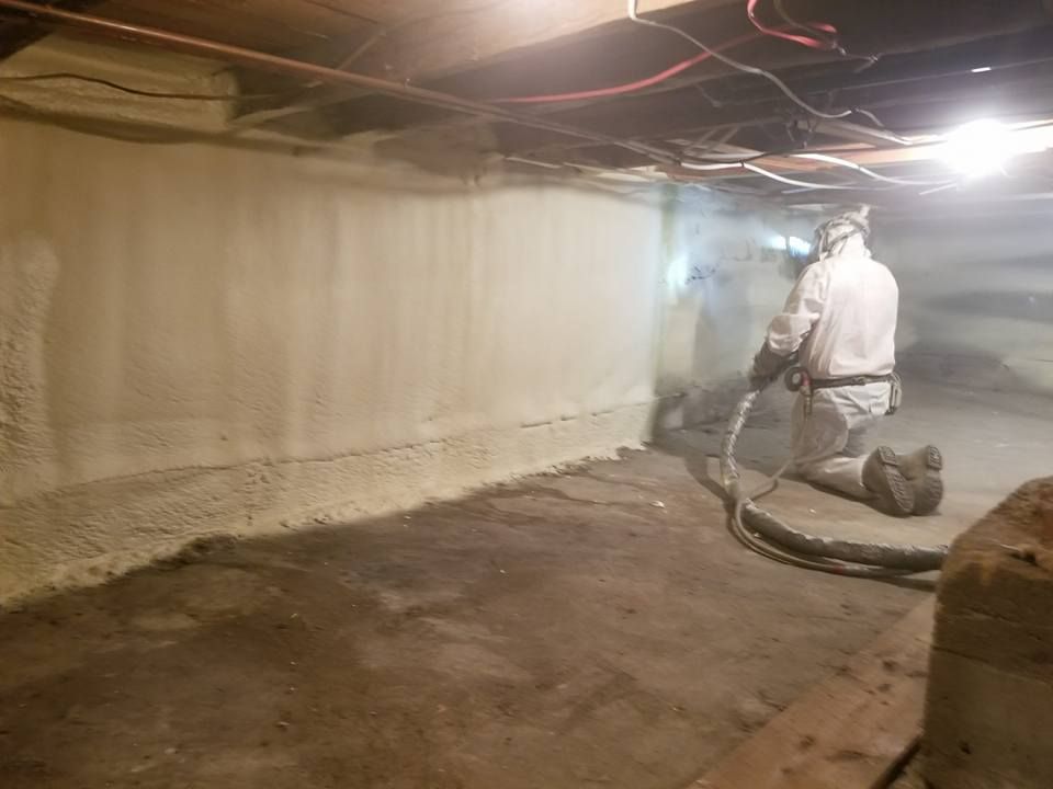 Une personne en combinaison de protection pulvérise de la mousse isolante dans un vide sanitaire.