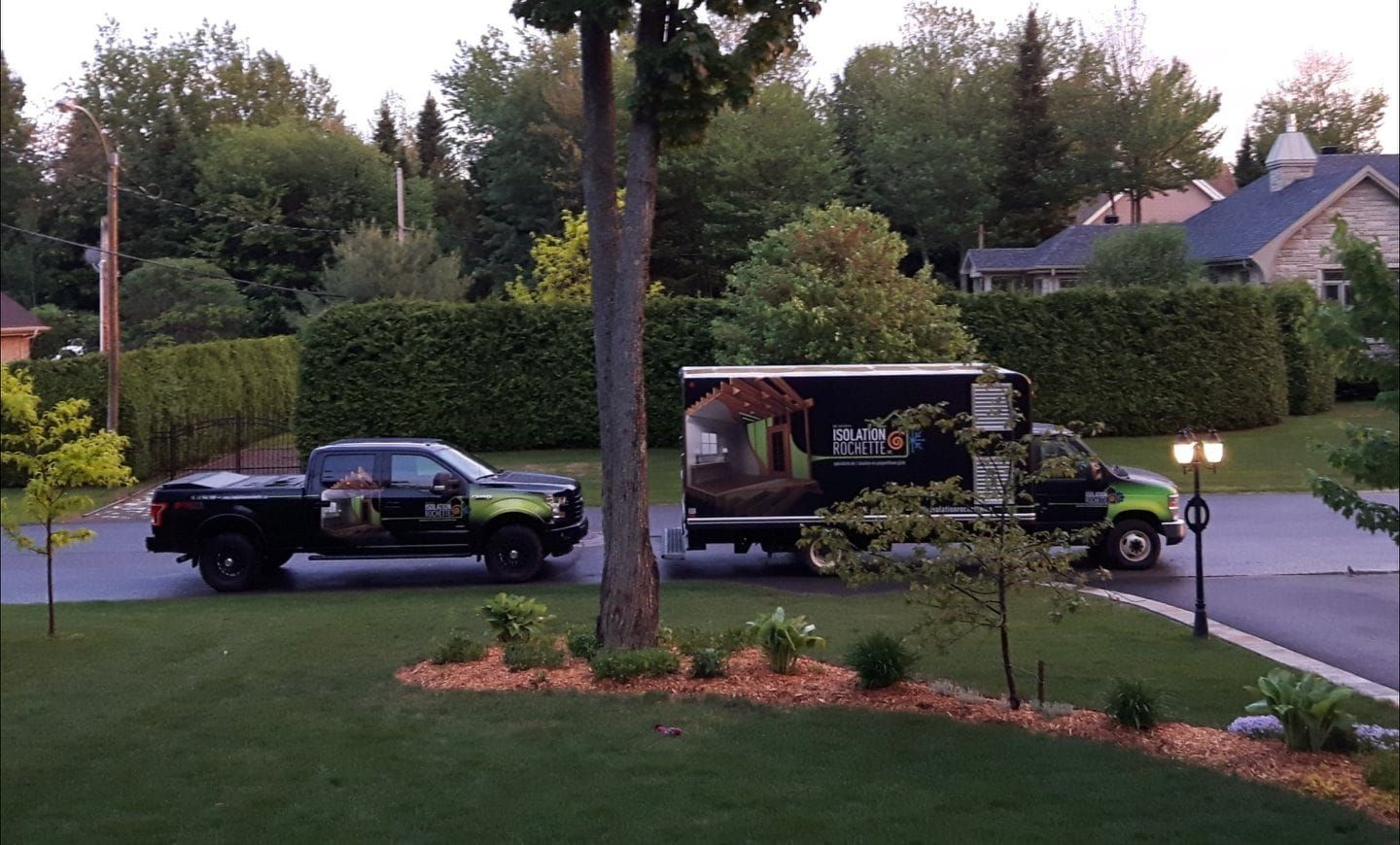 Un camion et une camionnette arborant des logos d'entreprise sont garés devant une maison. Pelouse verte, haie, arbre et maisons en arrière-plan.