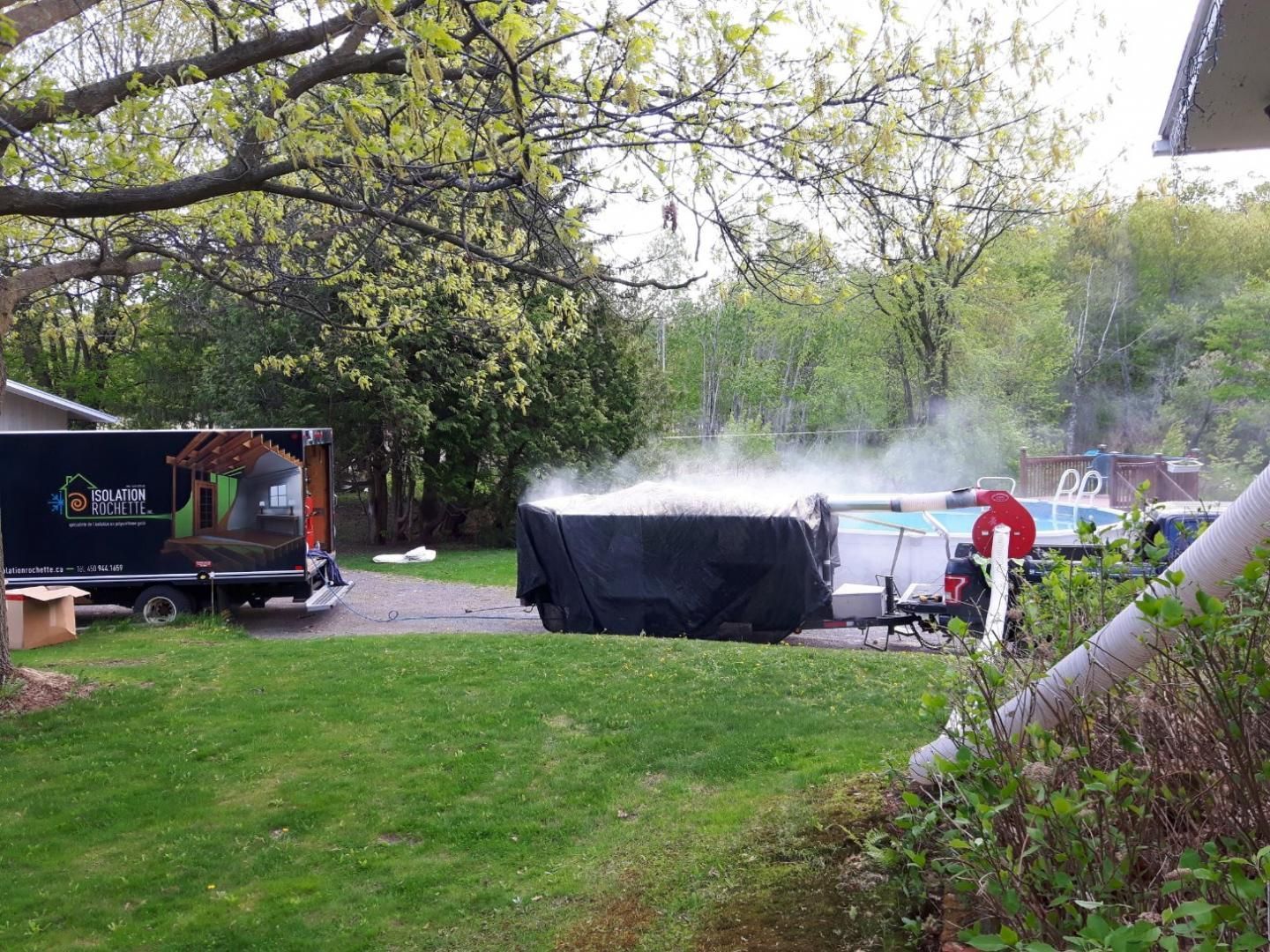 Une grande baignoire fumante recouverte d'une bâche noire, à côté d'une piscine et d'un camion de déménagement sur une pelouse.