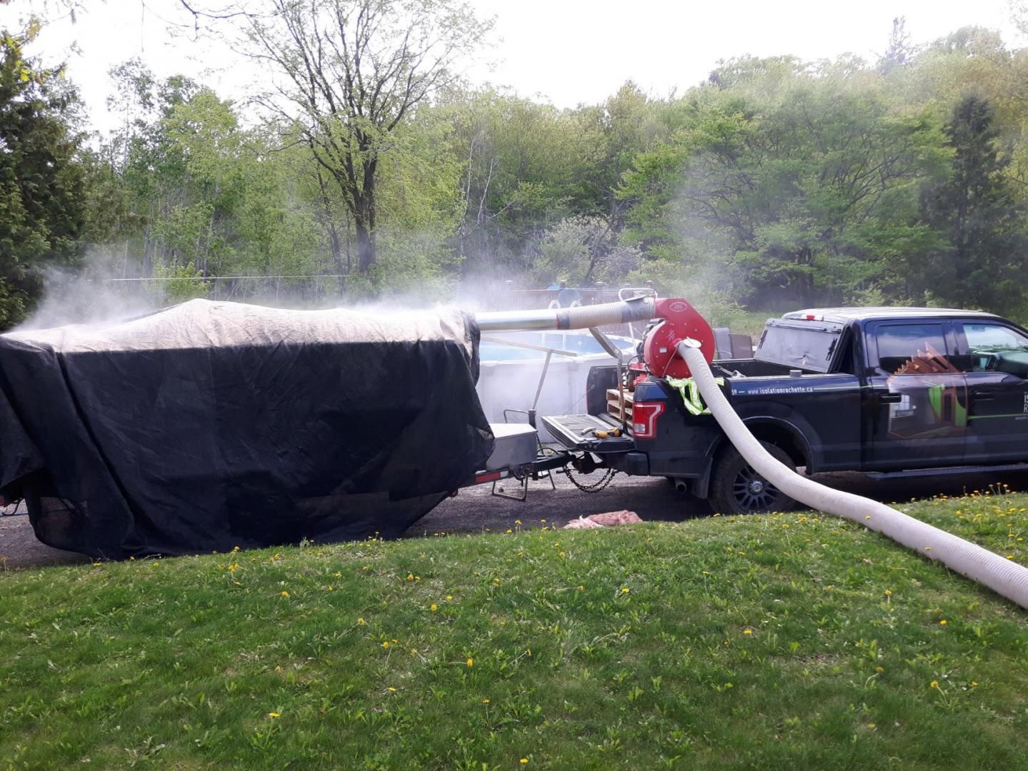 Un camion équipé d'un tuyau pompe du matériau dans une remorque couverte, créant un nuage de poussière blanche. Herbe verte et arbres.