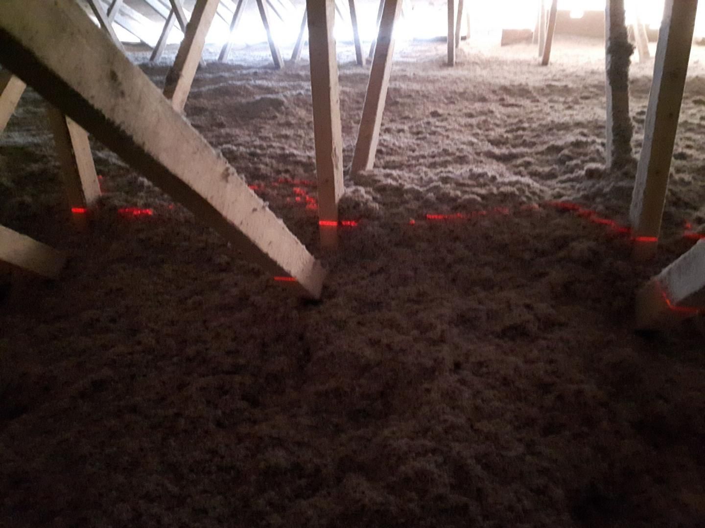 Isolation en cellulose brune dans un grenier, avec poutres en bois et lignes de nivellement laser rouges.