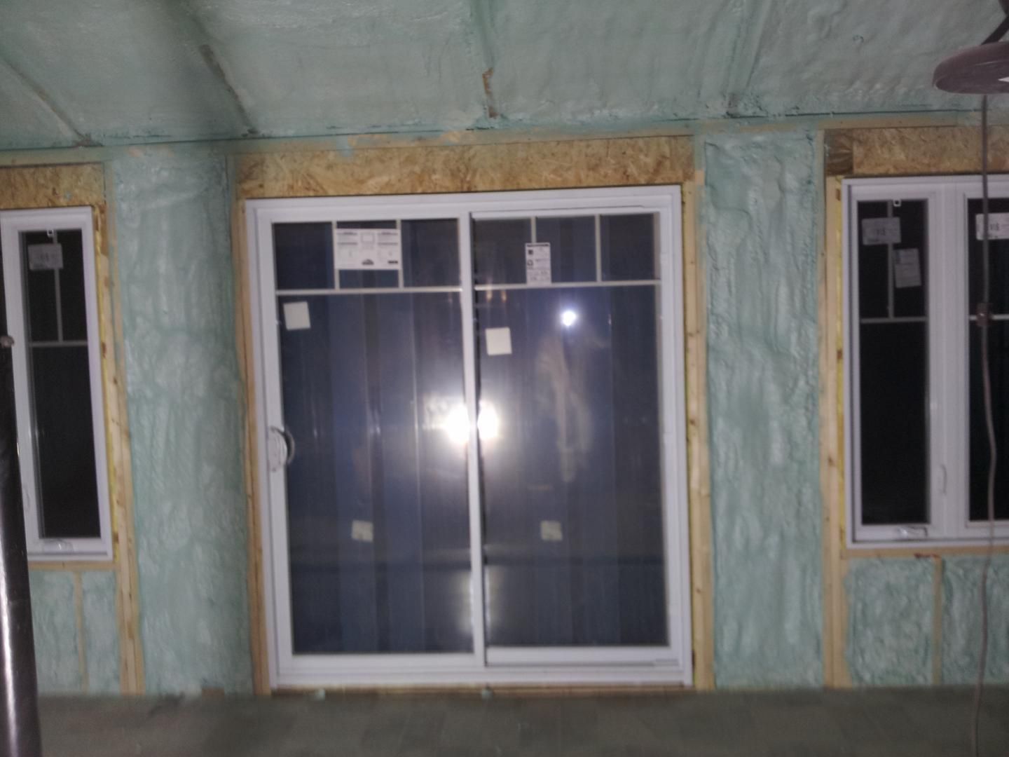 Vue intérieure d'une pièce en construction avec porte coulissante en verre et fenêtres encadrées de bois, isolées avec de la mousse bleu-vert.