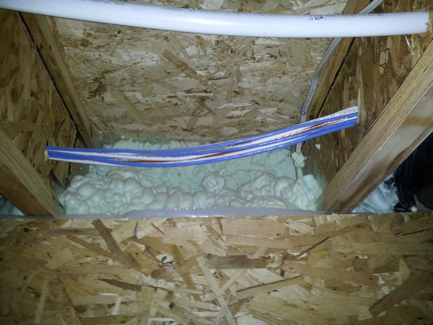 Isolation en mousse projetée entre des poutres en bois dans un grenier non aménagé, avec des fils électriques bleus et blancs et un tuyau blanc qui la traversent.