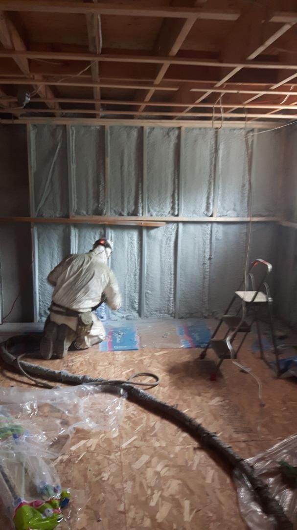 Une personne en combinaison de protection pulvérise de l'isolant dans une pièce. On aperçoit une structure en bois, un plancher en contreplaqué et une petite chaise.