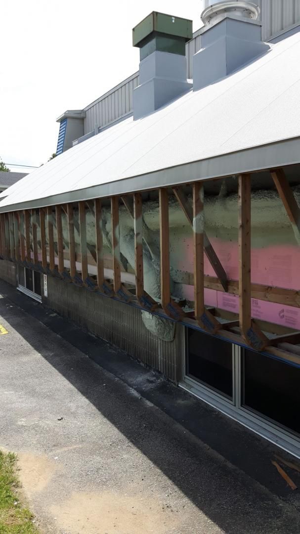 Extérieur du bâtiment en construction; charpente en bois apparente, isolation rose et fenêtres visibles.