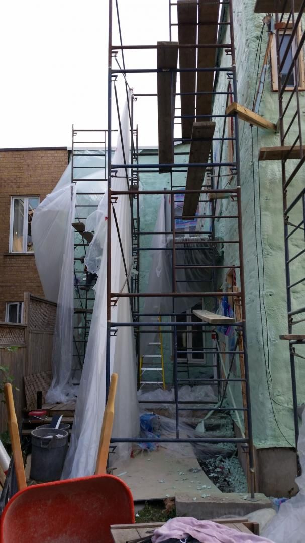 Échafaudage contre un bâtiment, avec des bâches en plastique, des planches de bois et une brouette rouge au premier plan.