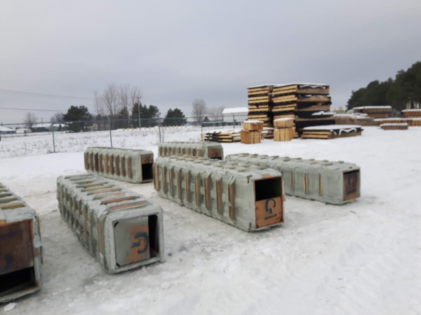 Des coffrages en béton sont posés dans une cour enneigée, avec des piles de bois et un ciel couvert en arrière-plan.