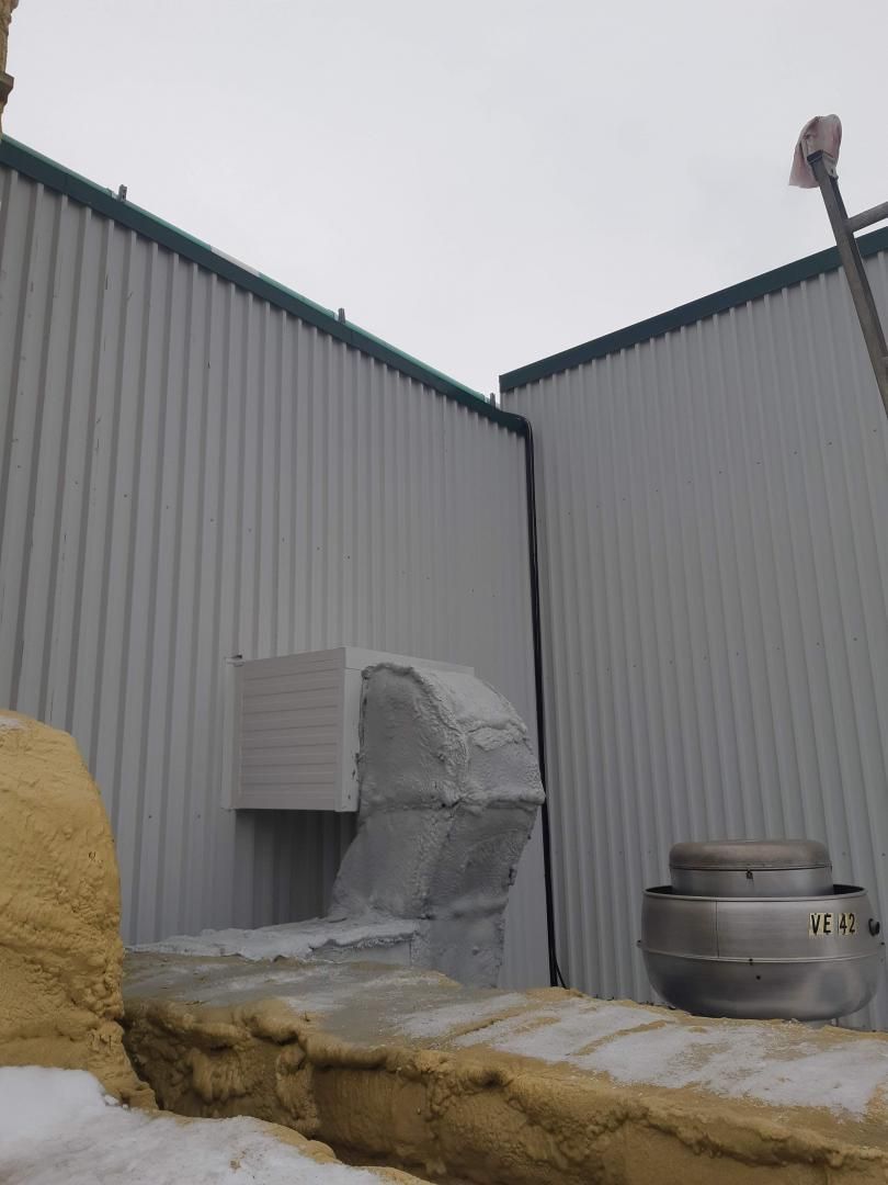 Extérieur d'un bâtiment avec une grande ventilation blanche recouverte d'un isolant gris clair. Sol enneigé.