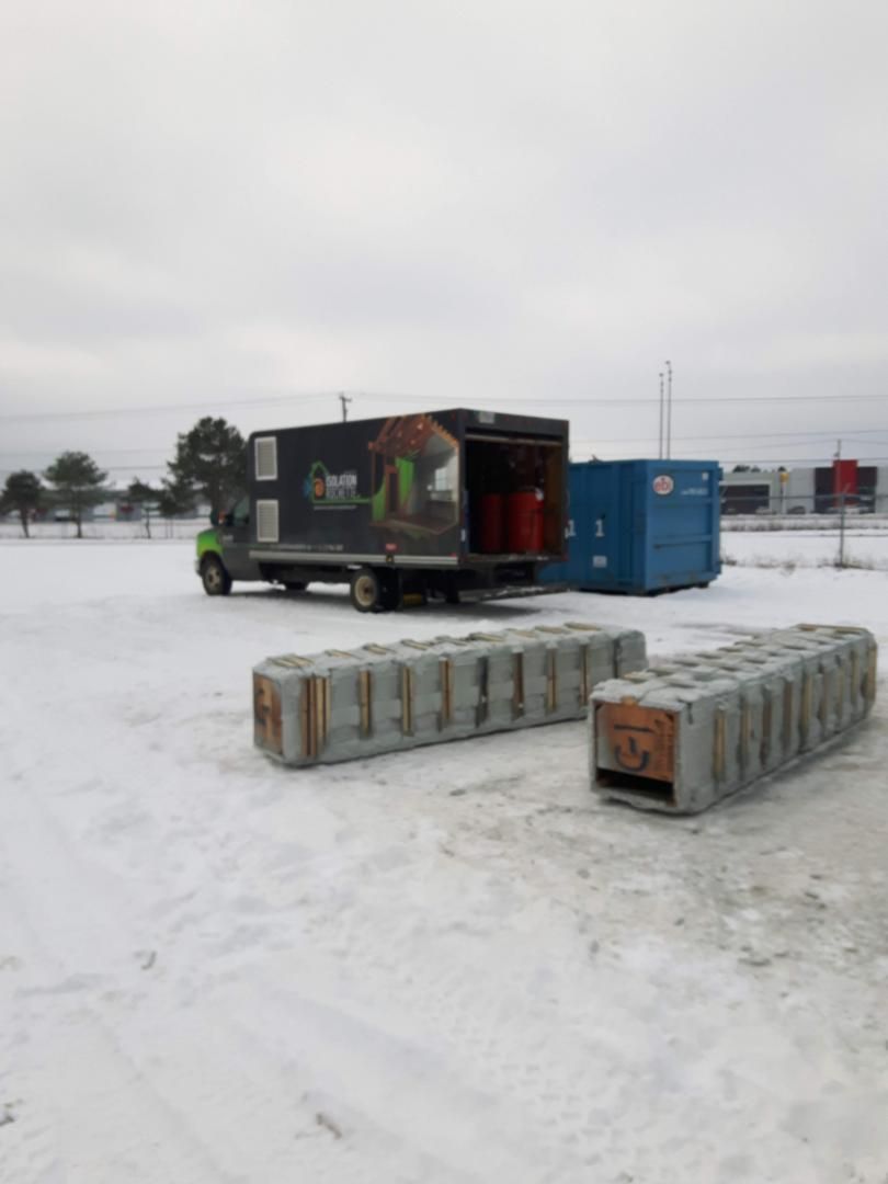 Un camion vert garé dans la neige, avec deux blocs de béton devant. Un conteneur bleu se trouve à côté du camion.