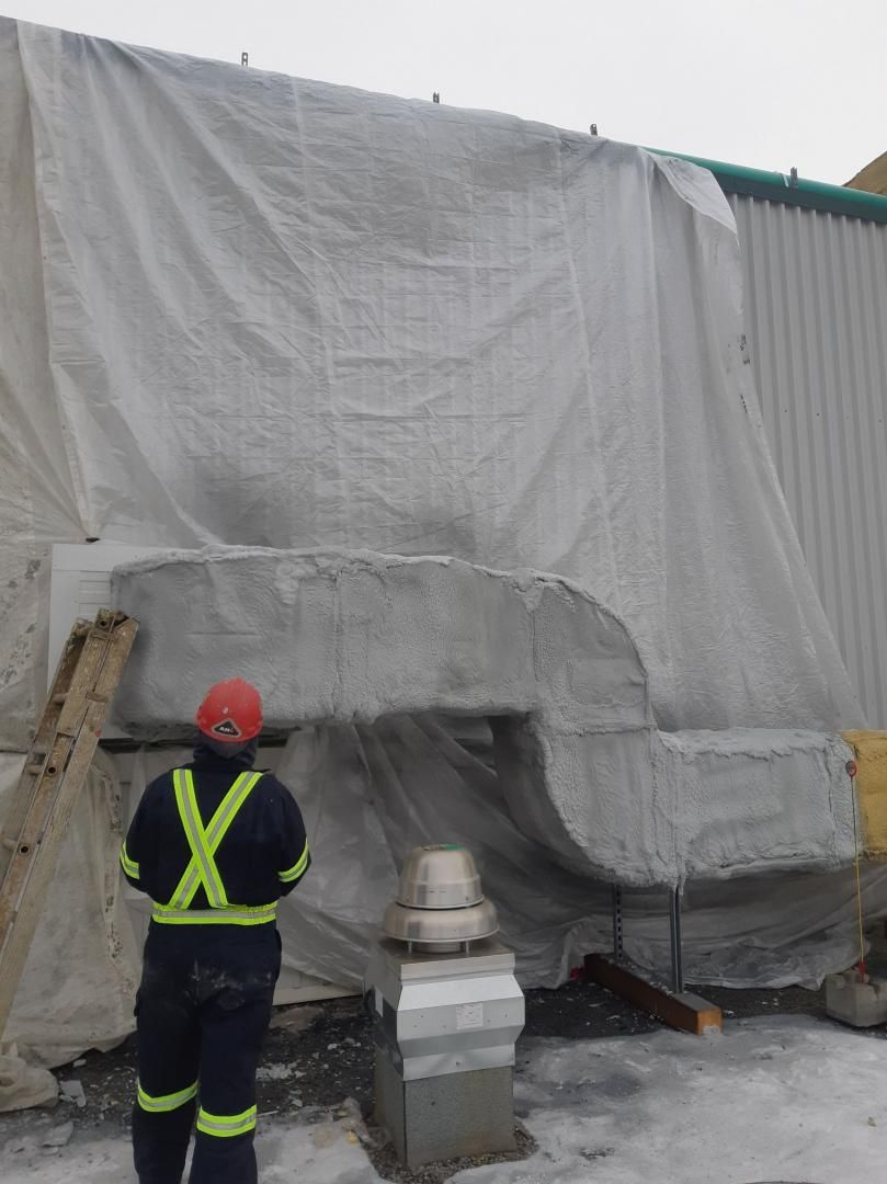Un ouvrier portant un gilet de sécurité et un casque observe une structure en béton recouverte d'une bâche en plastique près d'un bâtiment.