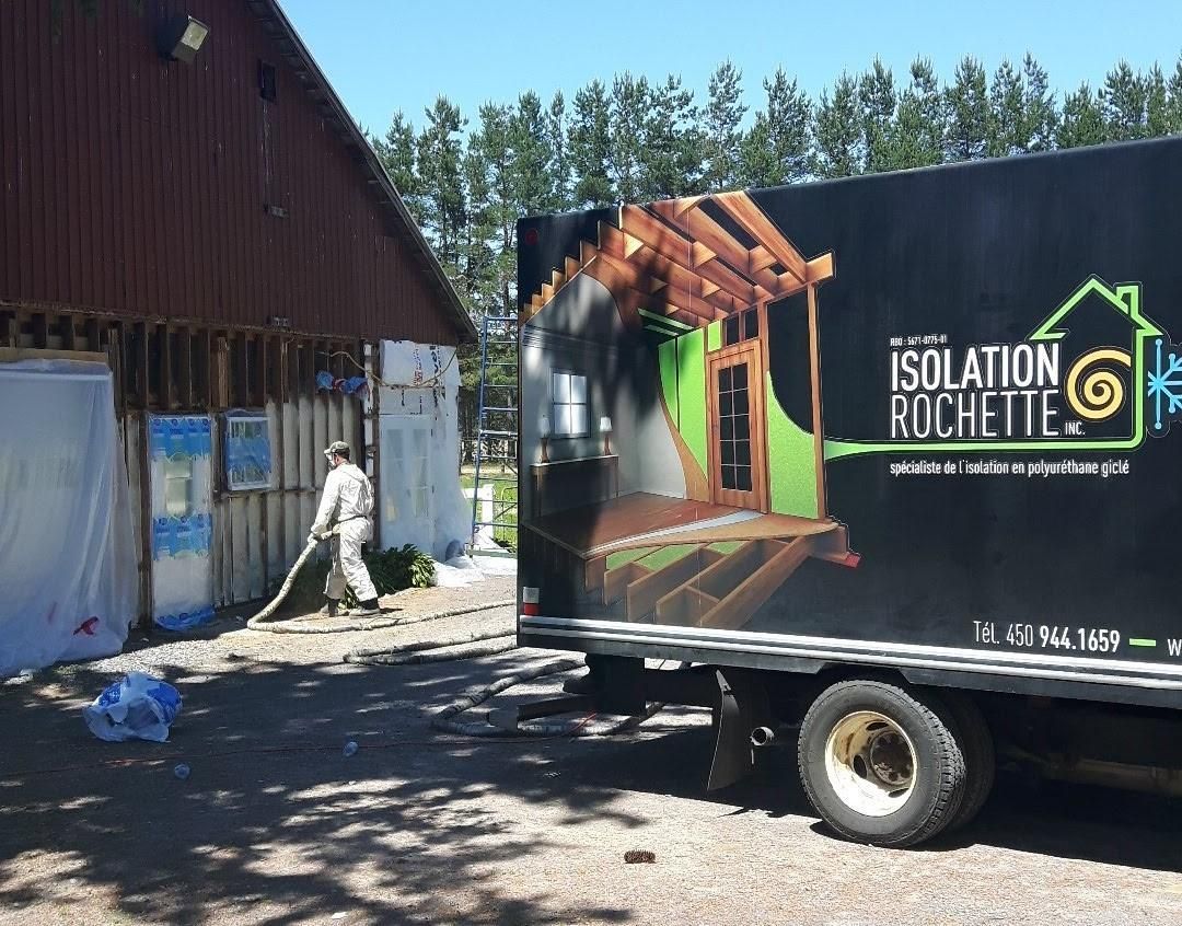 Camion avec le logo « Isolation Rochette » stationné près d'un bâtiment; un ouvrier nettoie les débris d'isolation.
