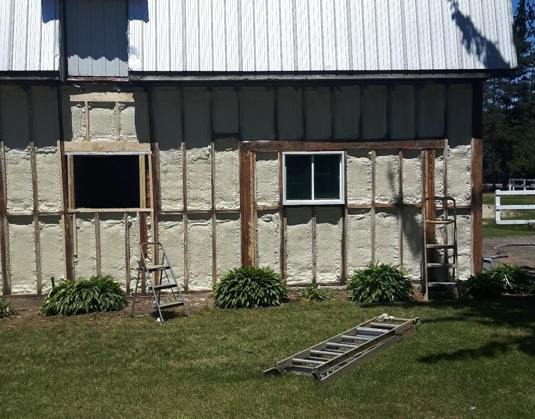 Extérieur de la grange avec isolation en mousse pulvérisée entre les montants, les fenêtres, l'échelle et les arbustes.