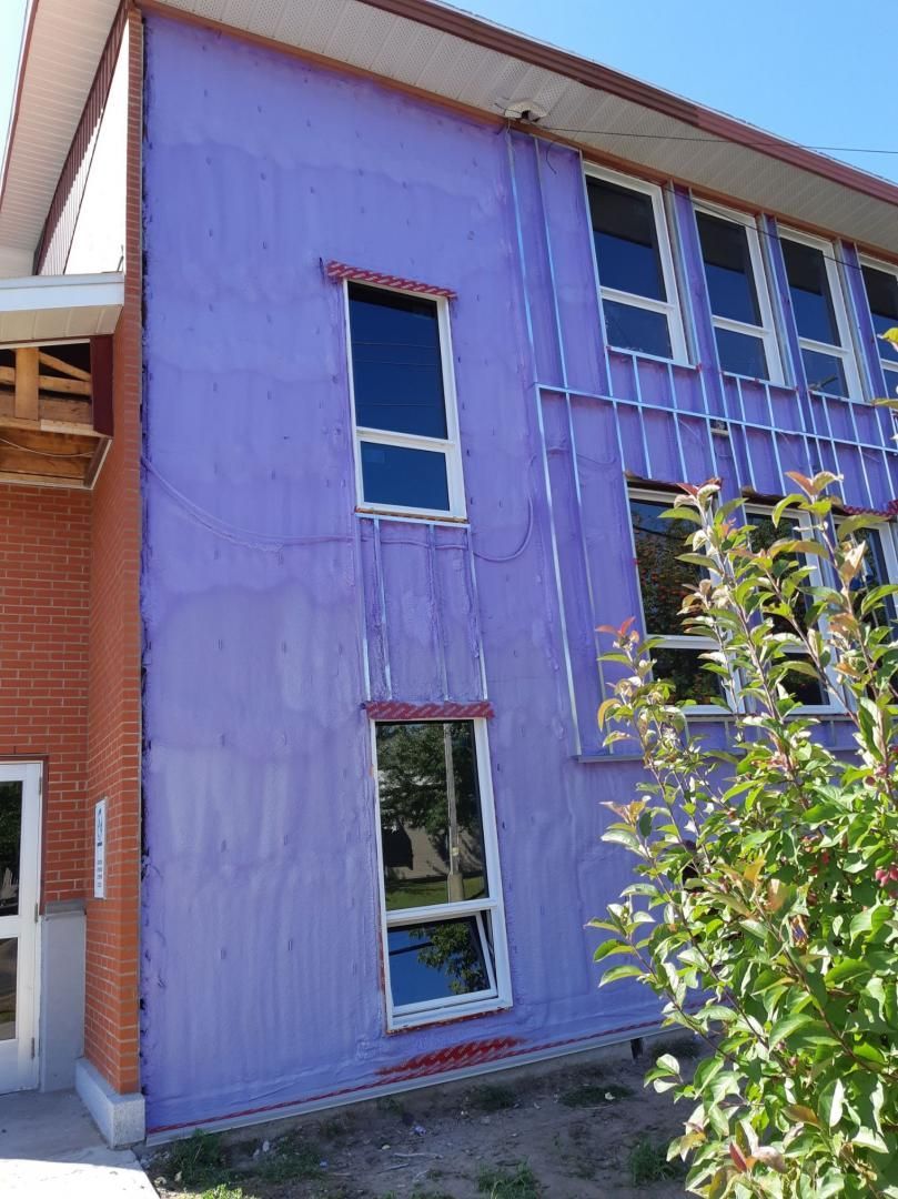 Bâtiment aux côtés violets avec plusieurs fenêtres ; des briques rouges et un arbre sont également visibles.
