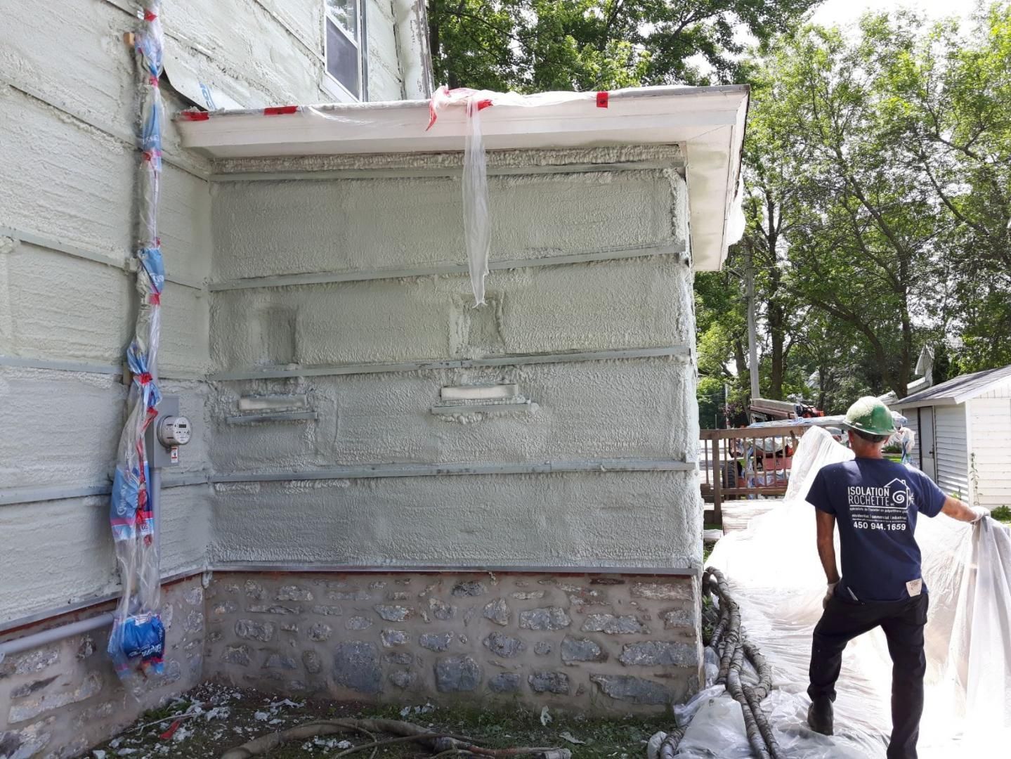 Un ouvrier du bâtiment près d'un bâtiment en cours d'isolation. De la mousse vert clair recouvre les murs, les fondations en béton et le jardin est arboré.