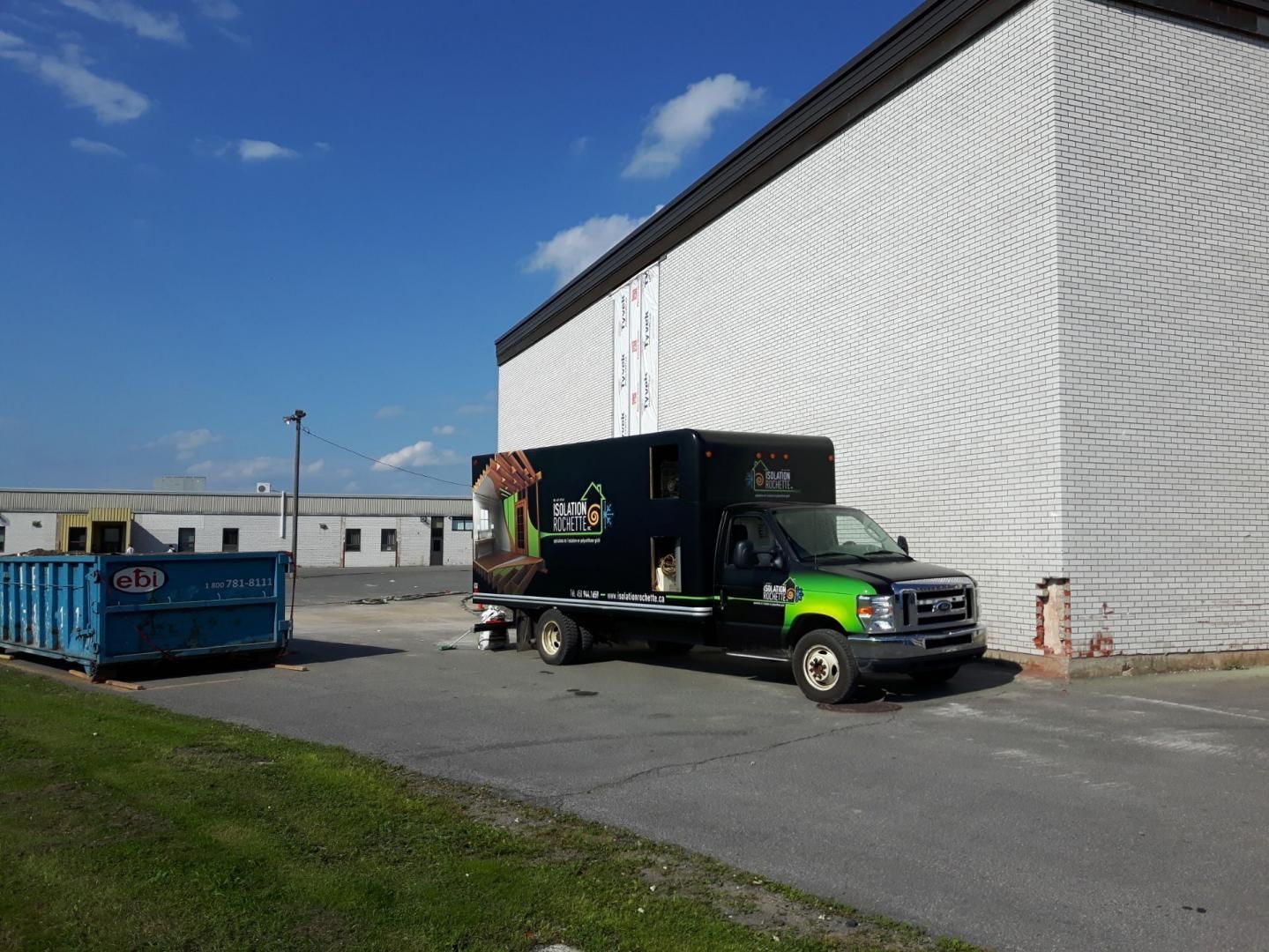 Camion avec logo garé près d'un bâtiment et d'une benne à ordures par une journée ensoleillée.