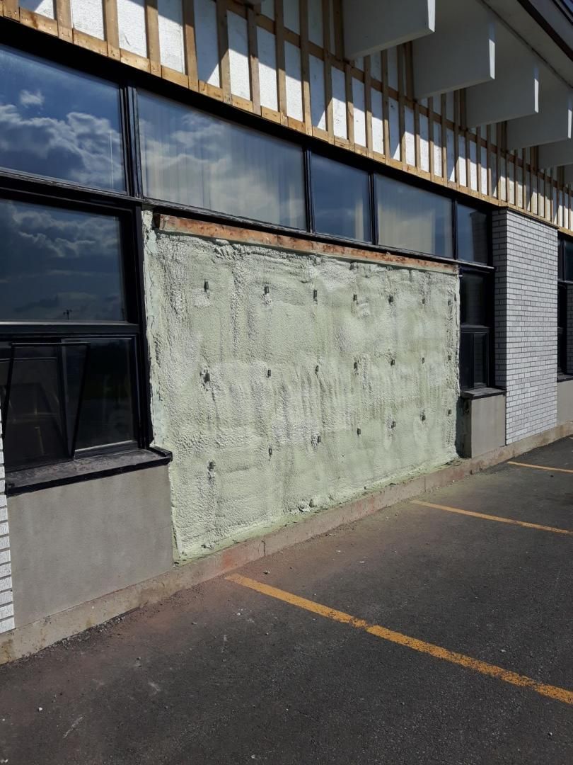 Bâtiment extérieur avec fenêtre barricadée ; isolation verte visible, cadres de fenêtres foncés, contours de voitures garées sur l'asphalte.