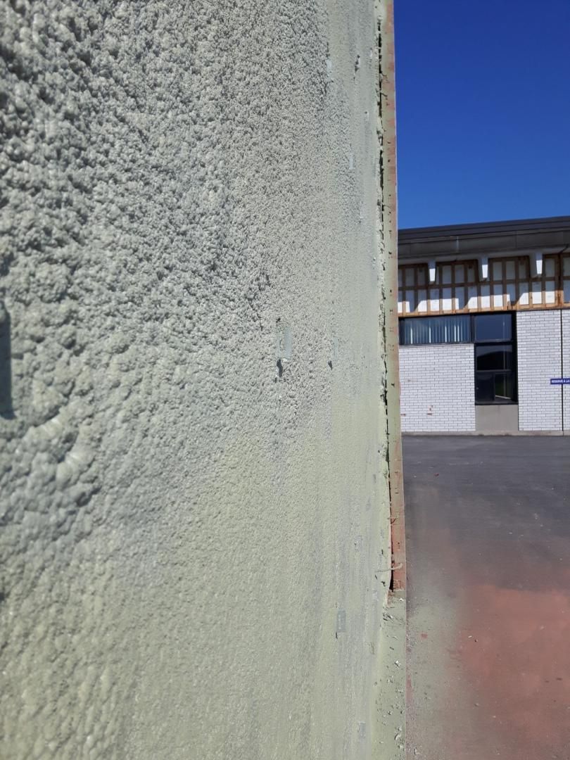 Mur texturé vert clair à côté d'une surface rouge, avec un bâtiment en arrière-plan sous un ciel bleu.