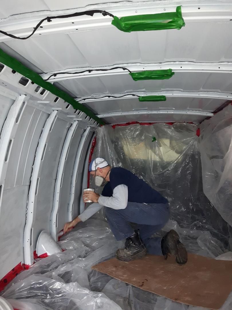 Personne peignant à l'intérieur d'une camionnette blanche, utilisant un pinceau, portant un masque et agenouillée sur du carton.